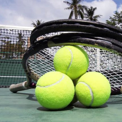 Three bright yellow tennis balls are stacked on an outdoor tennis court next to two tennis rackets, with trees and a net in the background—perfect for those seeking fun things to do in Costa Mesa.