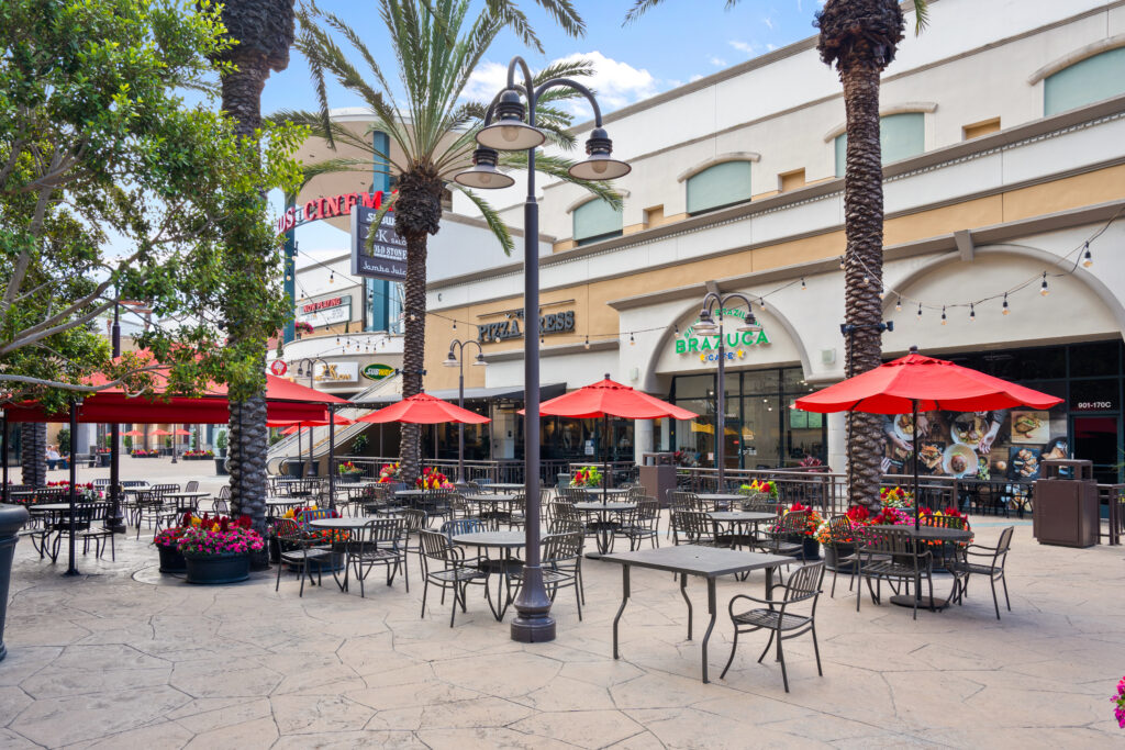 Outdoor dining area with empty tables and chairs under red umbrellas, surrounded by palm trees and colorful flower planters, set in front of restaurant storefronts on a sunny day in the vibrant Theater Arts District Costa Mesa.