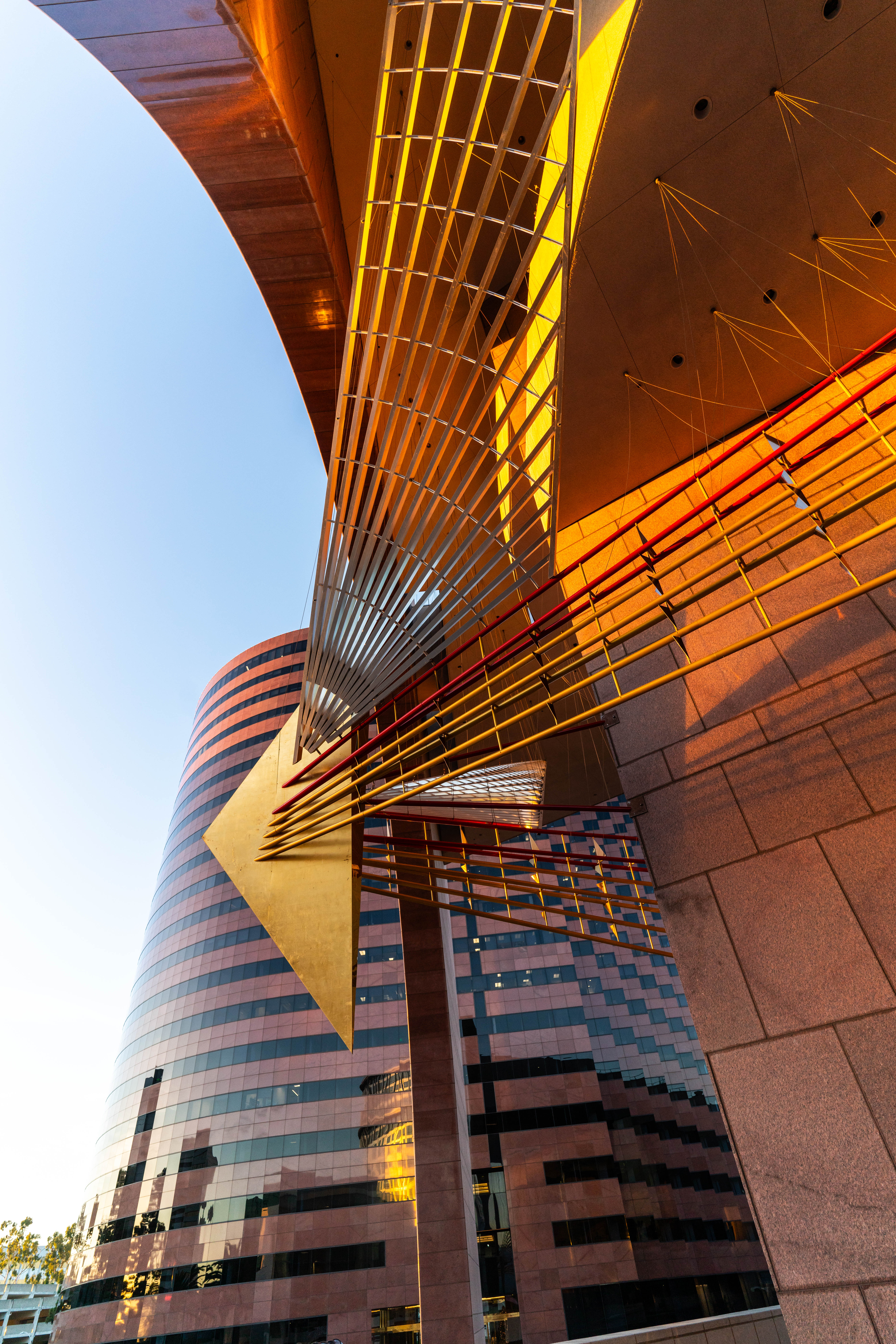 Modern architectural detail with gold and metallic abstract structures overhead, set against a curved glass office building reflecting the blue sky and sunlight—reminiscent of installations found along the Costa Mesa Art Walk.