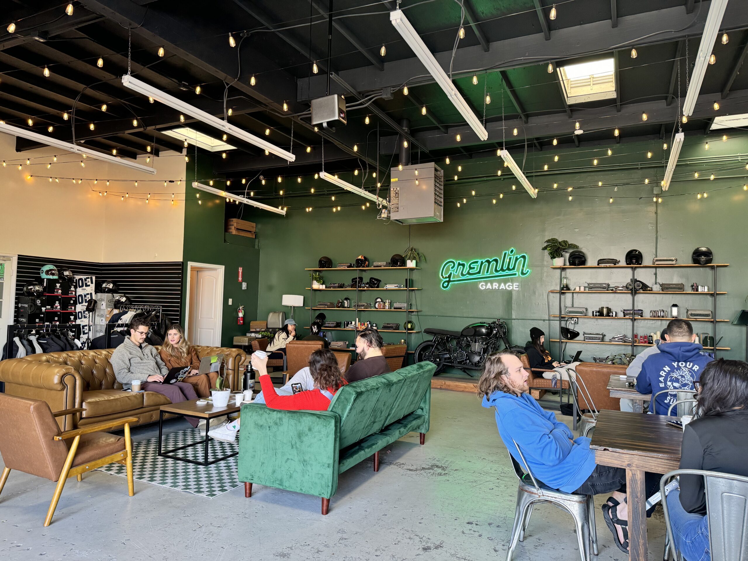 A modern lounge in Costa Mesa with green walls, leather and velvet sofas, people socializing and working at tables over coffee. Motorcycle helmets line the shelves beneath a neon “Gremlin Garage” sign, with string lights hanging from the ceiling.