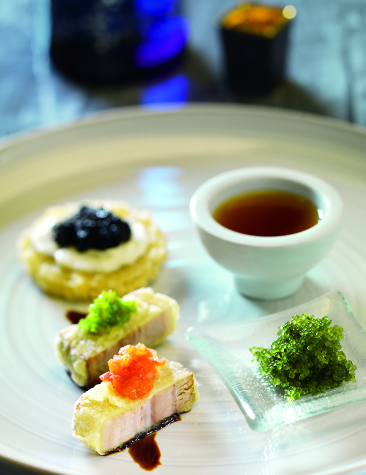 A gourmet dish featuring three pieces of seared fish topped with different caviars, a small cup of sauce, a crispy blini with black caviar, and green caviar—an artful creation inspired by the best sushi Costa Mesa has to offer.