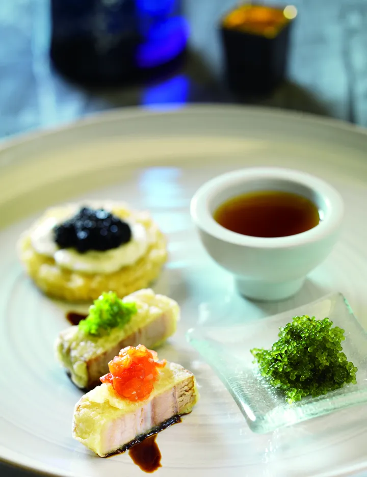 A gourmet dish featuring three pieces of seared fish topped with different caviars, a small cup of sauce, a crispy blini with black caviar, and green caviar—an artful creation inspired by the best sushi Costa Mesa has to offer.