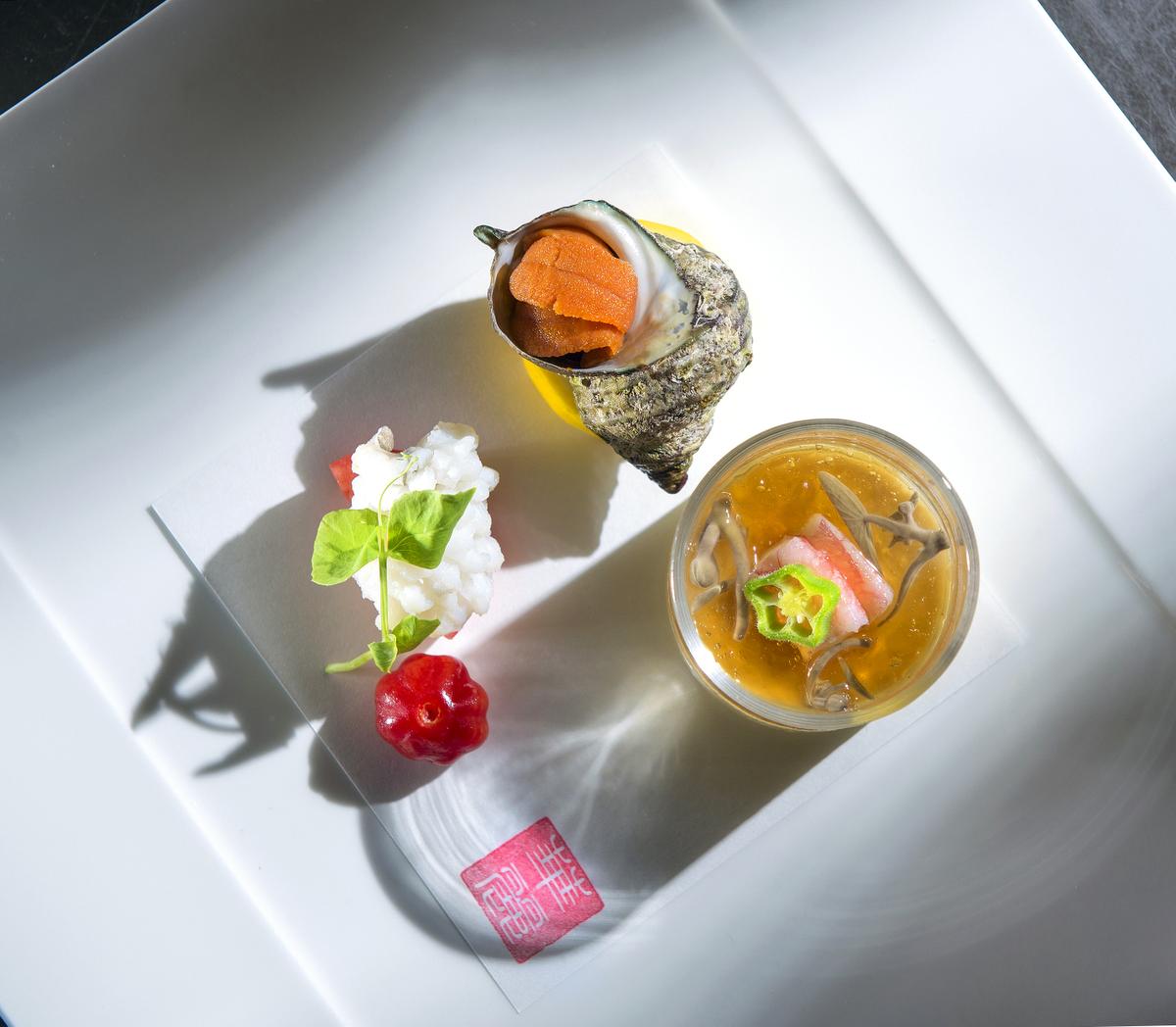 A square white plate with three artistic Japanese appetizers—showcasing the elegance you’d expect from the best sushi Costa Mesa offers: a seashell with orange garnish, a glass bowl of soup with vegetables, and white seafood topped with greens and a red flower-shaped item.