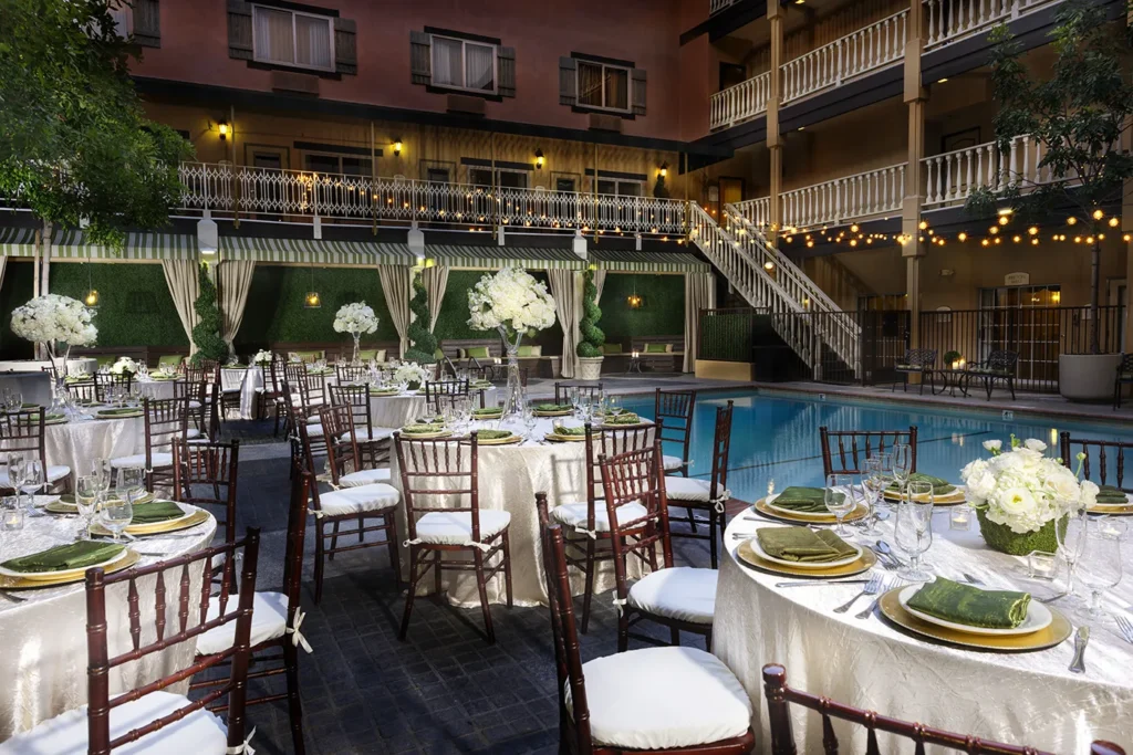 Elegant outdoor poolside event setup at one of the finest event venues in Costa Mesa, with round tables draped in white tablecloths, green napkins, and floral centerpieces. String lights glow overhead, with cabanas and a staircase completing the scene.