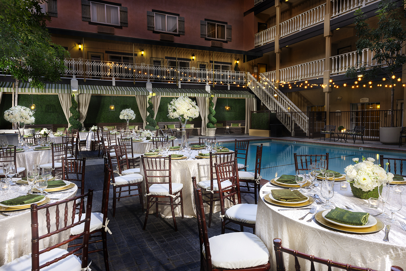 Elegant outdoor poolside event setup at one of the finest event venues in Costa Mesa, with round tables draped in white tablecloths, green napkins, and floral centerpieces. String lights glow overhead, with cabanas and a staircase completing the scene.