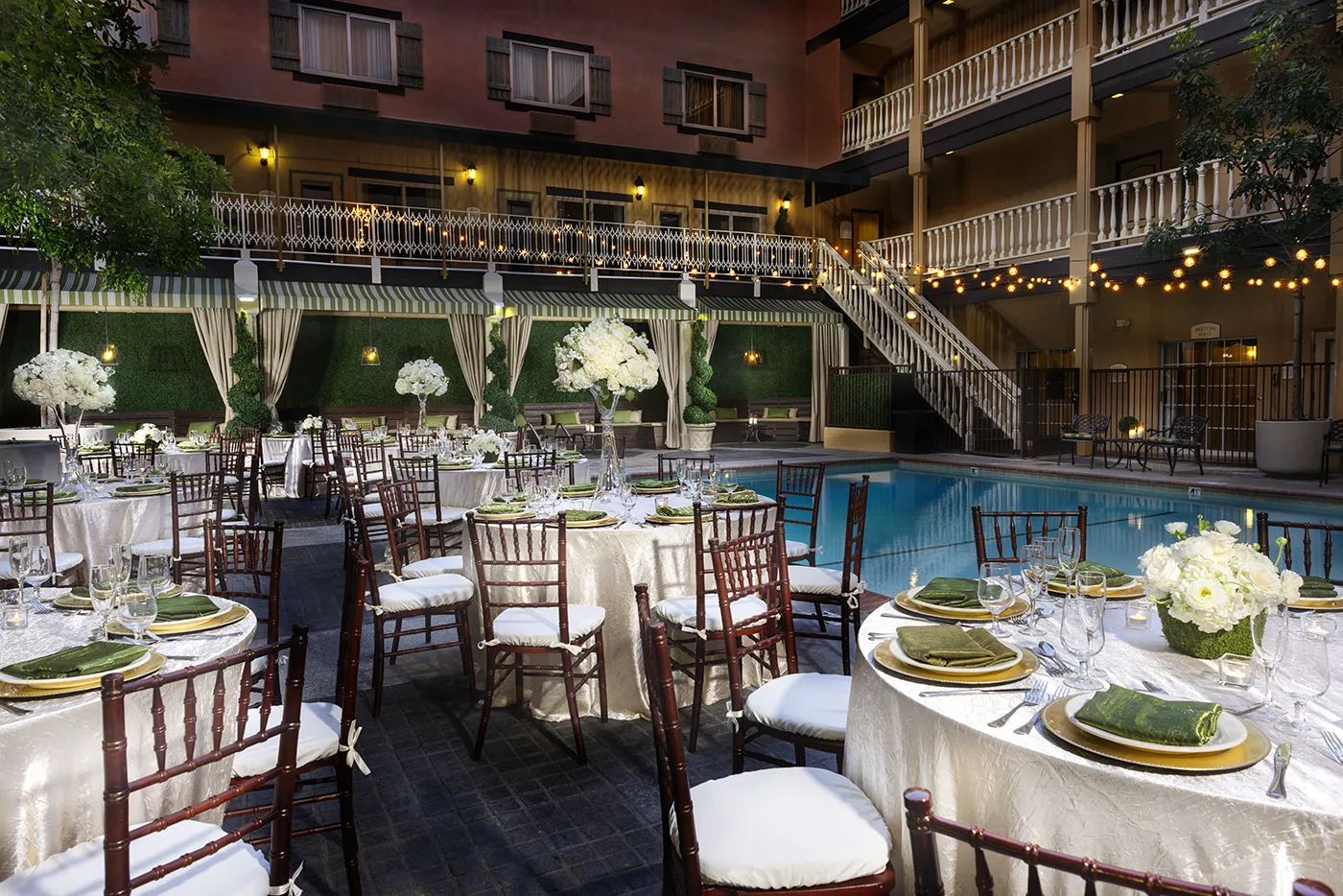Elegant outdoor poolside event setup at one of the finest event venues in Costa Mesa, with round tables draped in white tablecloths, green napkins, and floral centerpieces. String lights glow overhead, with cabanas and a staircase completing the scene.