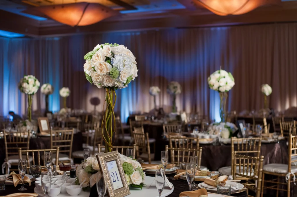 An elegant banquet hall among top wedding venues Costa Mesa, set for an event with round tables, gold chairs, tall white and pale blue floral centerpieces, framed table numbers, and softly lit draped walls in the background.