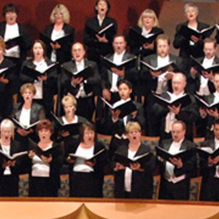 A choir of adults dressed in formal black and white attire sings from sheet music on stage at the Segerstrom Performing Arts Center Costa Mesa. Their mouths are open mid-song, appearing focused and engaged.