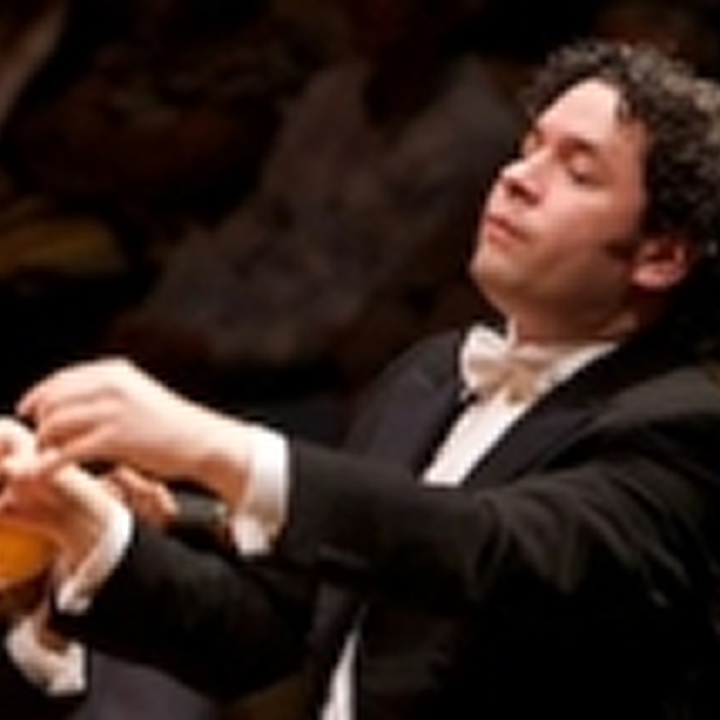 A man in a black suit and white bow tie conducts music with expressive hand gestures, set against a blurred, dark background at the Segerstrom Performing Arts Center in Costa Mesa.