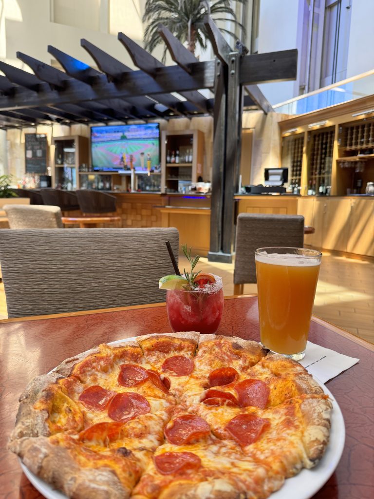 A pepperoni pizza sits on a table beside a pint of beer and a red cocktail with a lime garnish. In the background, there is a bar area, lounge chairs, and a TV showing a sports game.