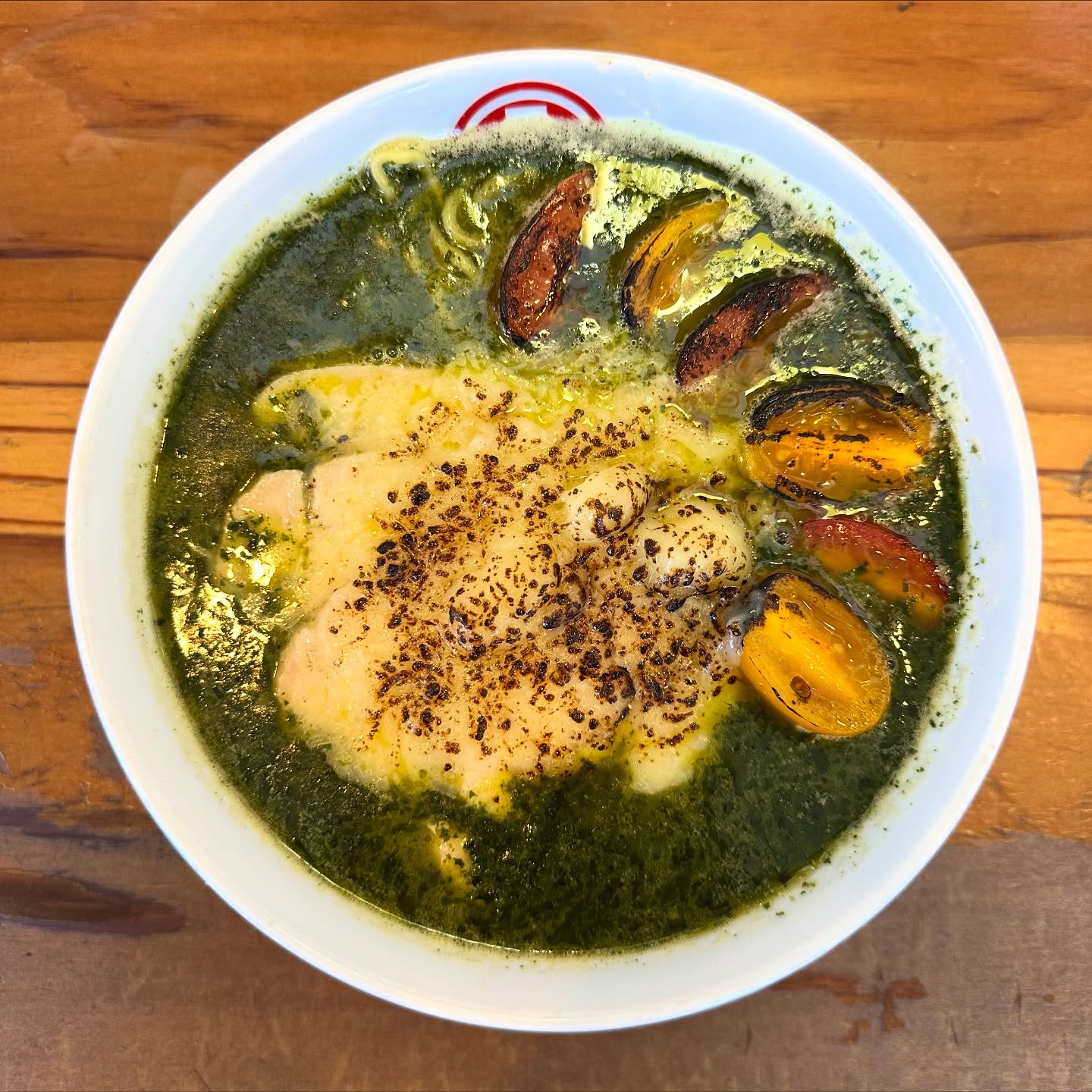 A bowl of ramen with green broth, grilled cherry tomatoes, a slice of roasted chicken, and a sprinkle of black pepper on top, served on a wooden table.