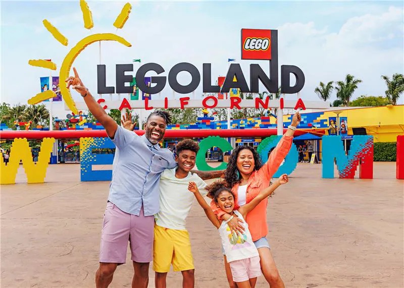 A smiling family of four poses with arms raised in front of the colorful LEGOLAND California entrance sign, a perfect destination for day trips from Costa Mesa, with large LEGO blocks and a bright welcome display in the background.