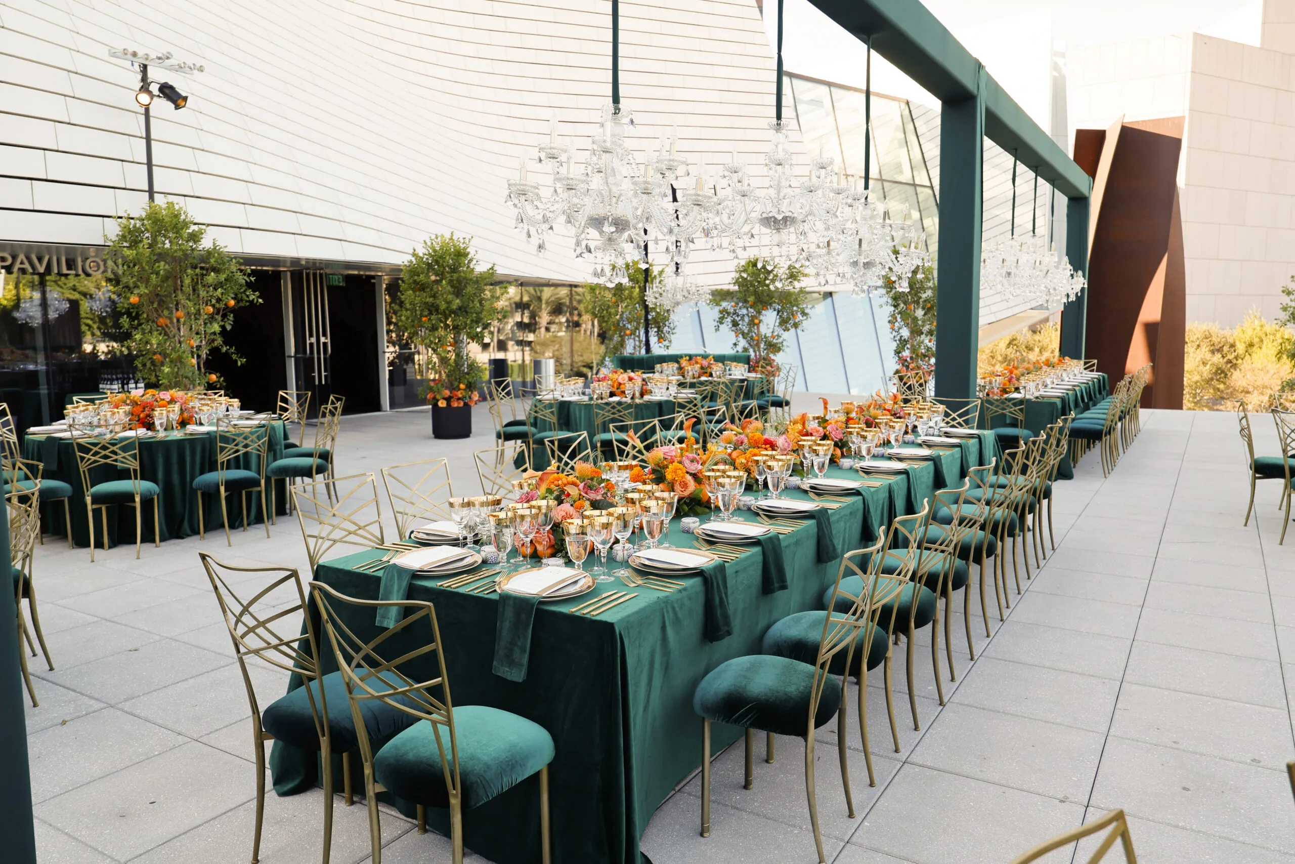 Elegant outdoor dining setup with long tables covered in green velvet tablecloths, gold chairs, vibrant orange floral centerpieces, glassware, and chandeliers overhead—an ideal scene for event venues in Costa Mesa, surrounded by modern architecture and greenery.