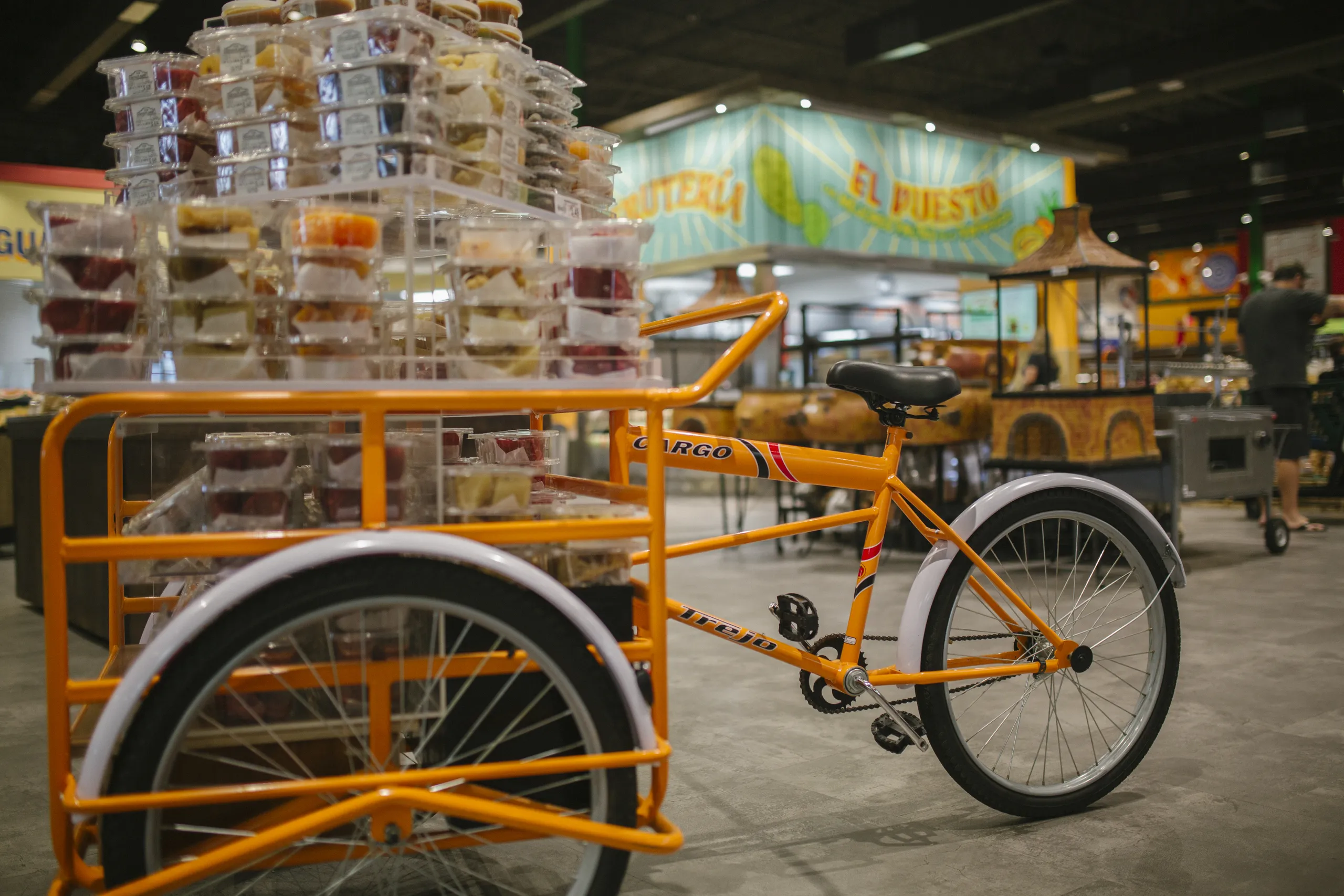 An orange cargo tricycle carries stacked plastic containers of food in a bustling Costa Mesa, California market setting, with colorful signs and vibrant displays in the background.