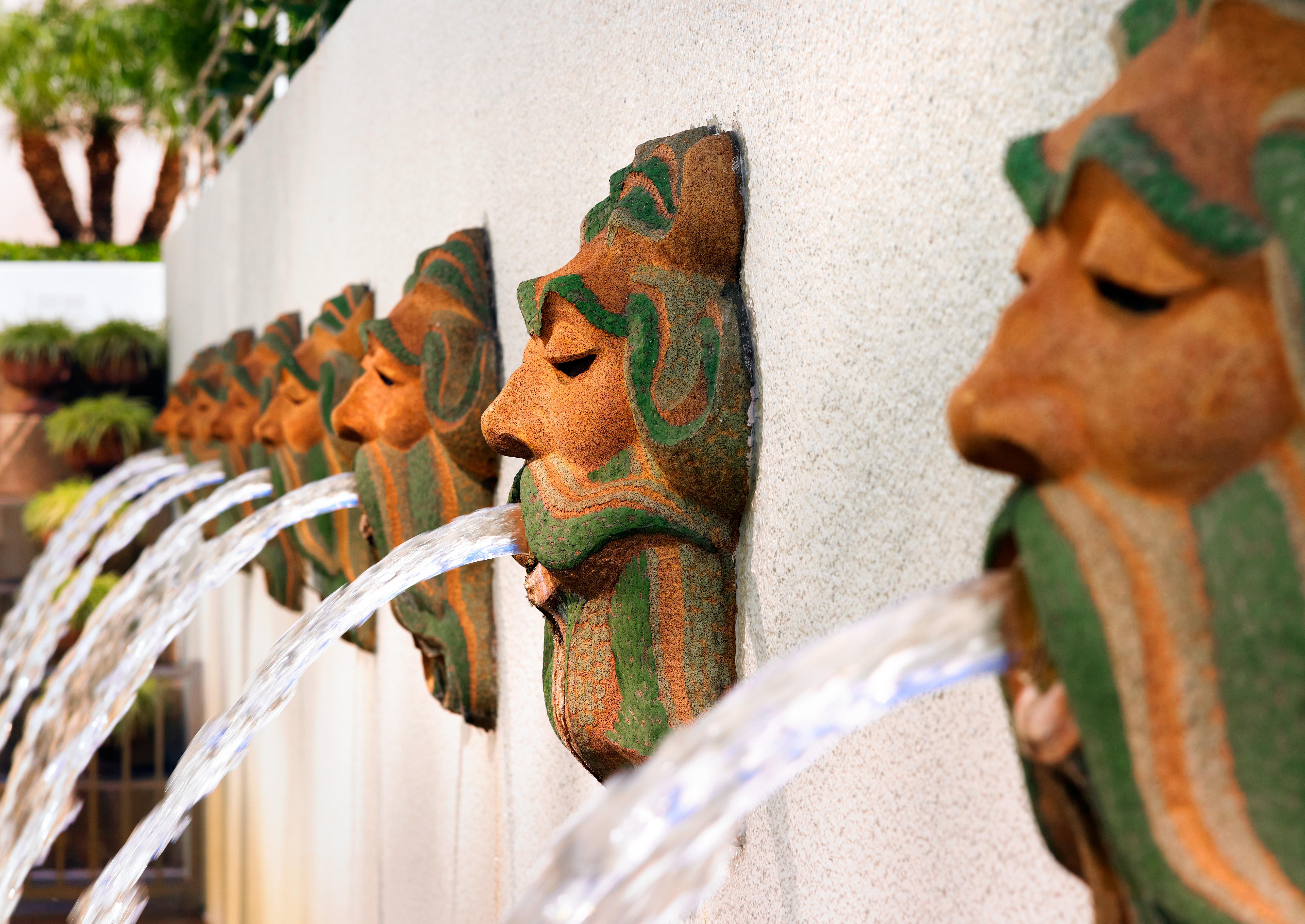 Bronze-green decorative face sculptures on a stone wall spout streams of water into a fountain, creating a repeating pattern—an inviting scene reminiscent of the creative spirit found along the Costa Mesa Art Walk. Lush greenery completes the backdrop.