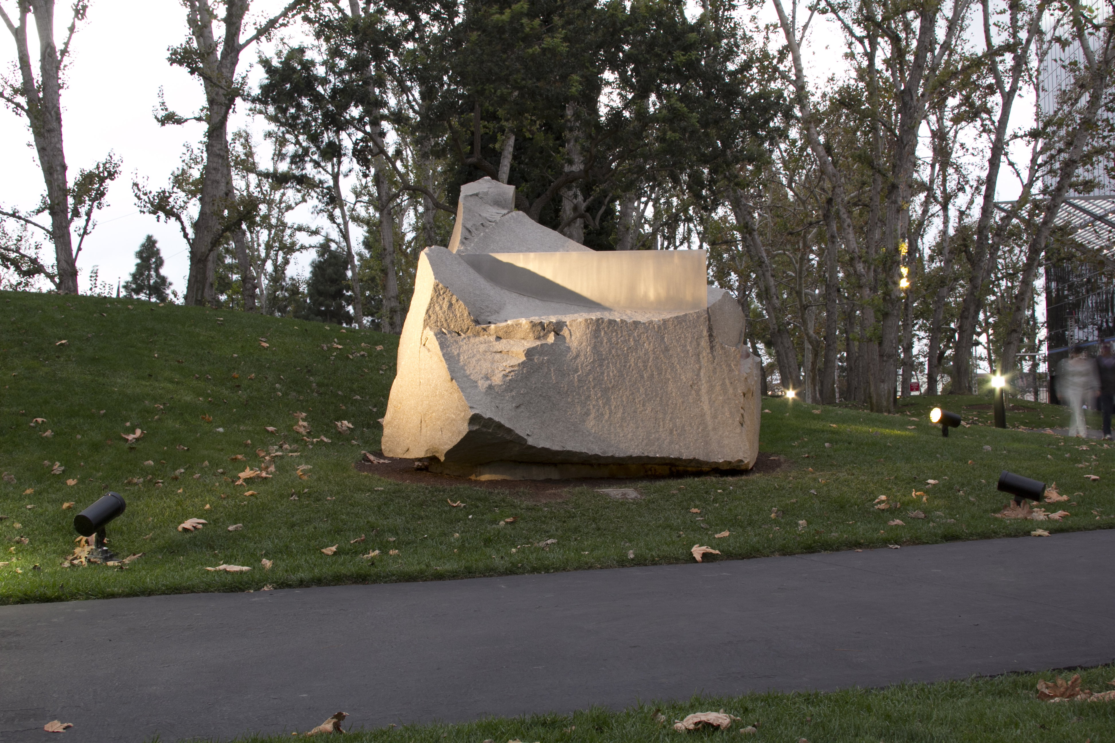 A large, rough-hewn stone sculpture sits on a grassy hill surrounded by trees, illuminated by spotlights. Along the Costa Mesa Art Walk, a paved pathway winds past fallen leaves scattered on the grass.