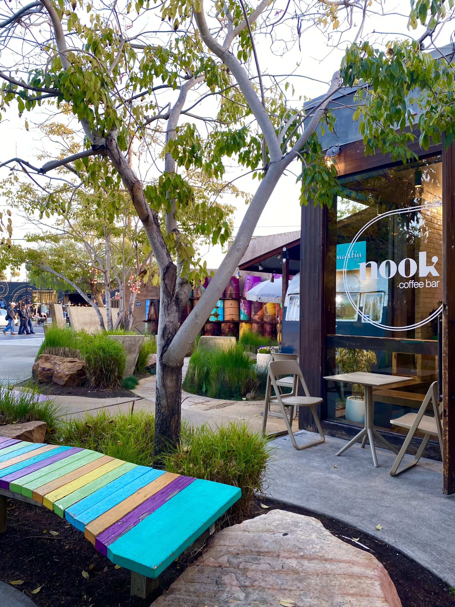 A colorful rainbow-painted bench sits under a leafy tree outside Nook Coffee Bar in Costa Mesa, where two wooden chairs and a small table invite coffee lovers to relax by large windows. Green plants and rocks decorate the cozy outdoor space.