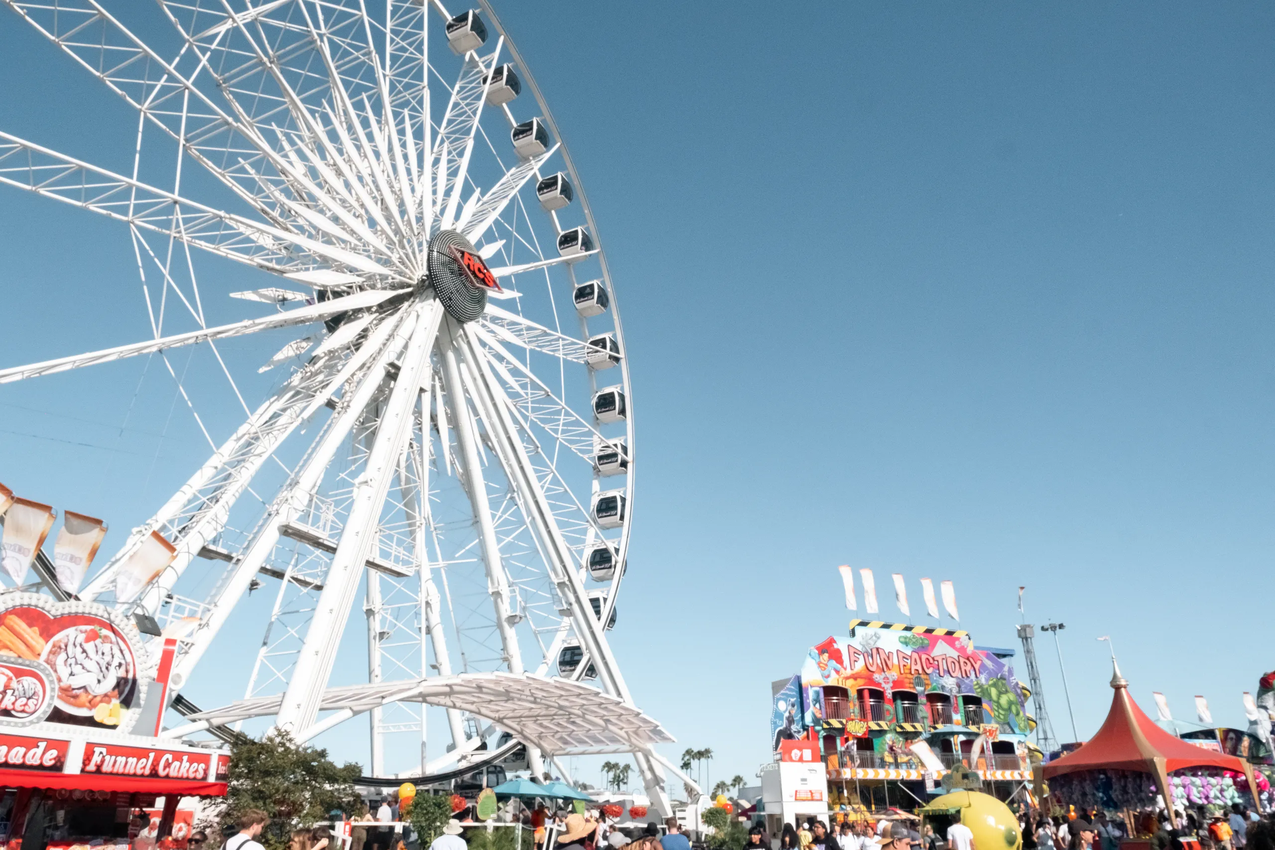 A large Ferris wheel towers over a busy fairground in Costa Mesa, California, with colorful booths, a funhouse, and a clear blue sky overhead. People are walking and enjoying the festivities.