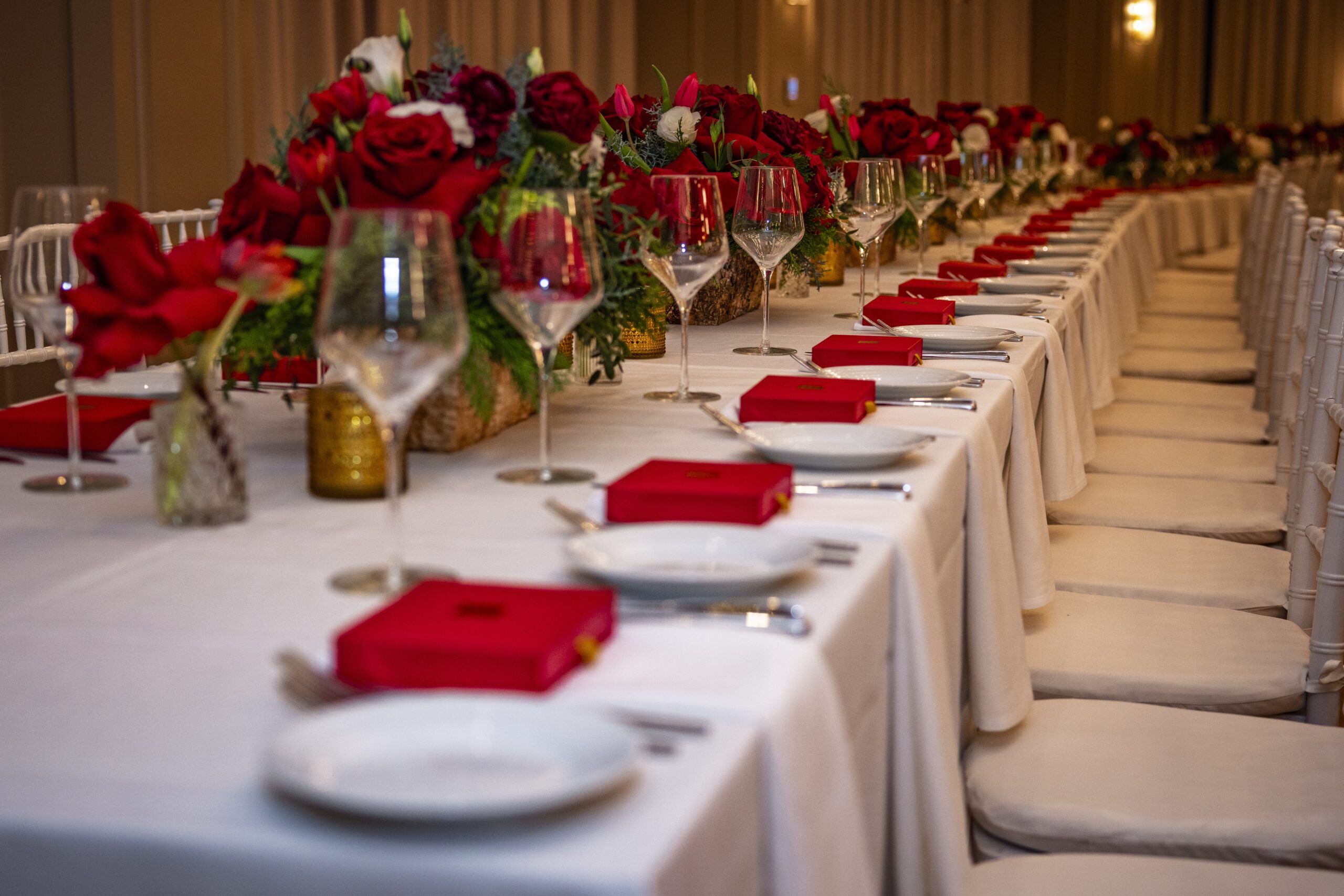 A long banquet table with white tablecloths is set with white plates, wine glasses, silverware, and red gift boxes—perfect for elegant wedding venues Costa Mesa. The centerpiece features red roses and greenery, with white chairs lining both sides.