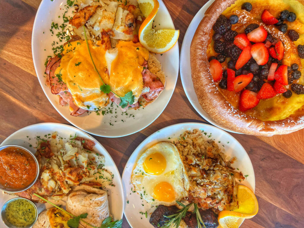 Four plates of breakfast food on a wooden table, featuring eggs Benedict with potatoes and oranges, a large Dutch baby pancake topped with berries, a plate with eggs and hash browns, and another plate with eggs, potatoes, and salsa.