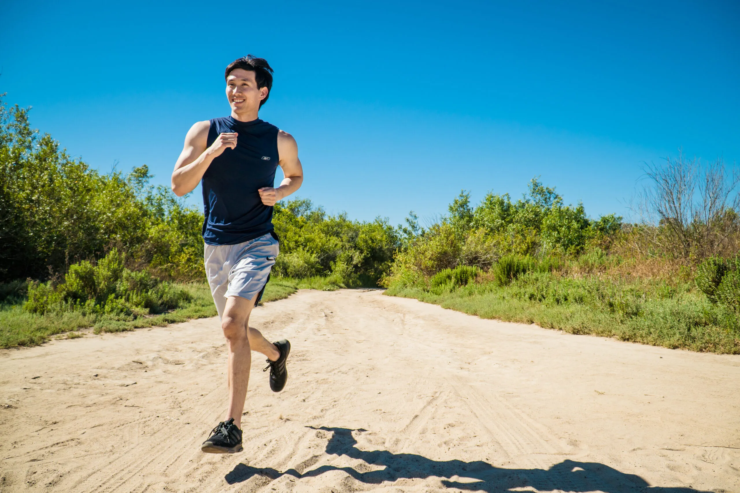 A man in a navy sleeveless shirt and gray shorts is jogging on a dirt trail near green bushes and trees under a clear blue sky, his route inspired by the scenic paths on the Costa Mesa map.