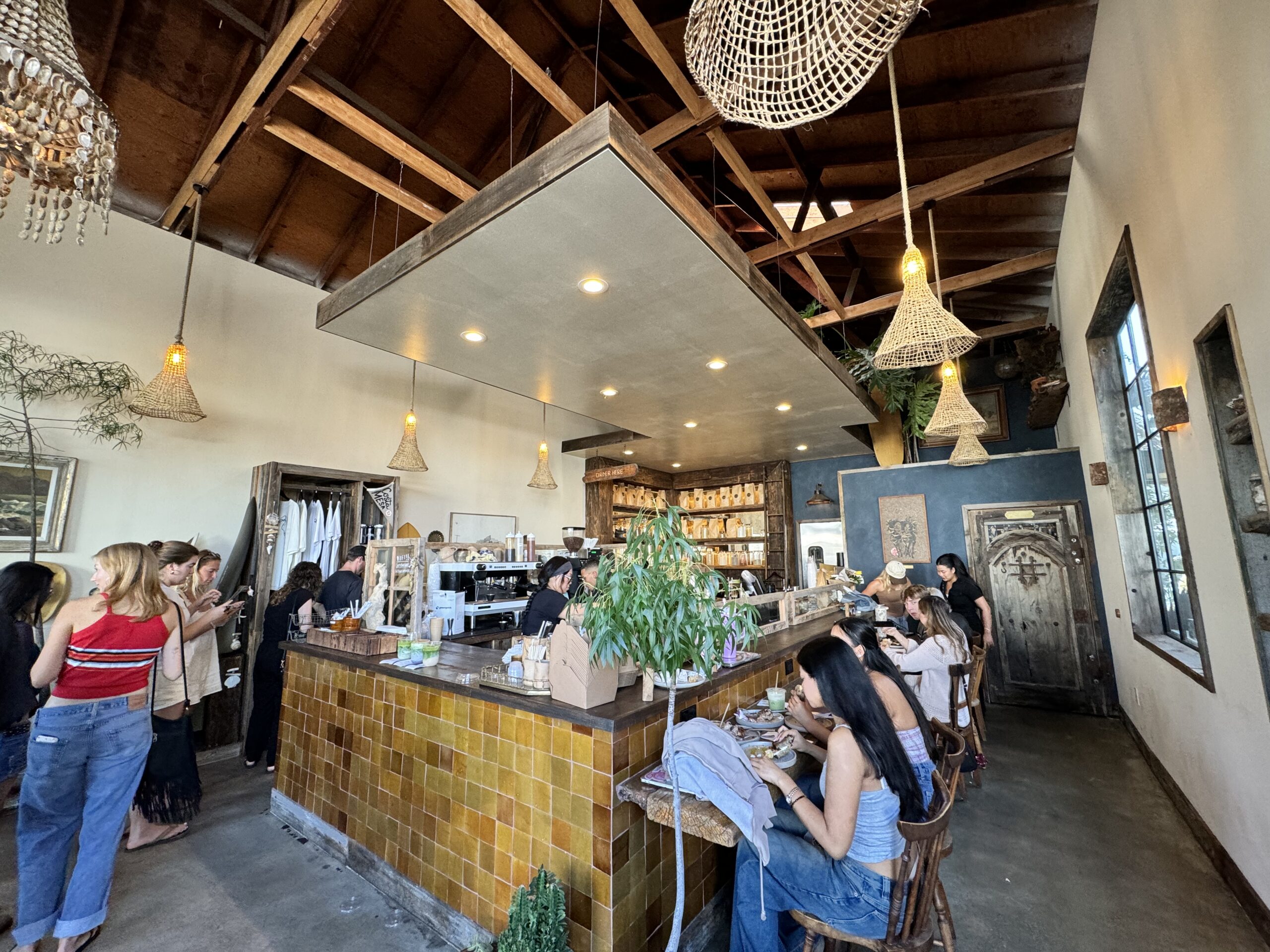 A cozy café offering the best brunch Costa Mesa has to offer, with people ordering at the counter and others relaxing at wooden tables. Warm lighting, hanging wicker lamps, plants, and rustic decor complement the high, exposed wood ceiling.