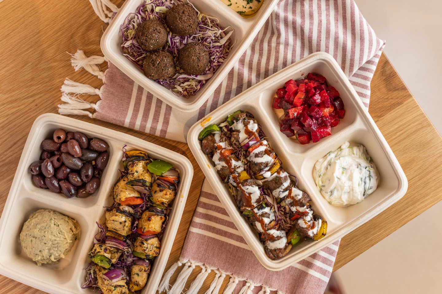 Three compostable takeout containers filled with Mediterranean food, including falafel, olives, grilled skewers, sauces, and vibrant salads, arranged on a striped cloth on a wooden table.