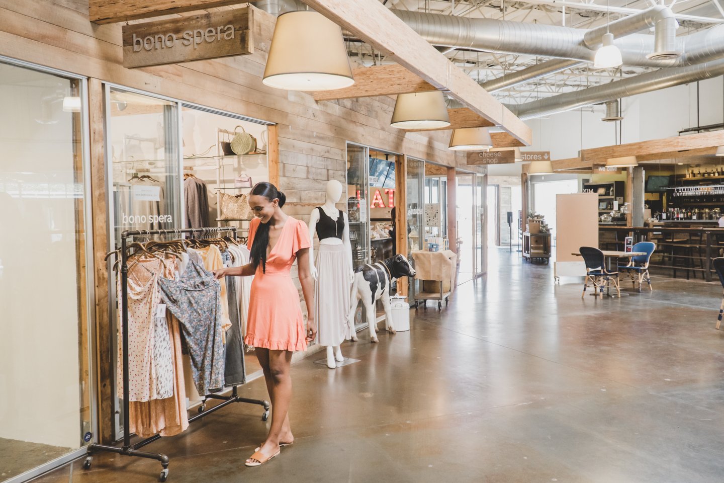A woman in a coral dress browses clothing on a rack inside a modern, spacious boutique with wooden accents and large glass store fronts under industrial lighting.
