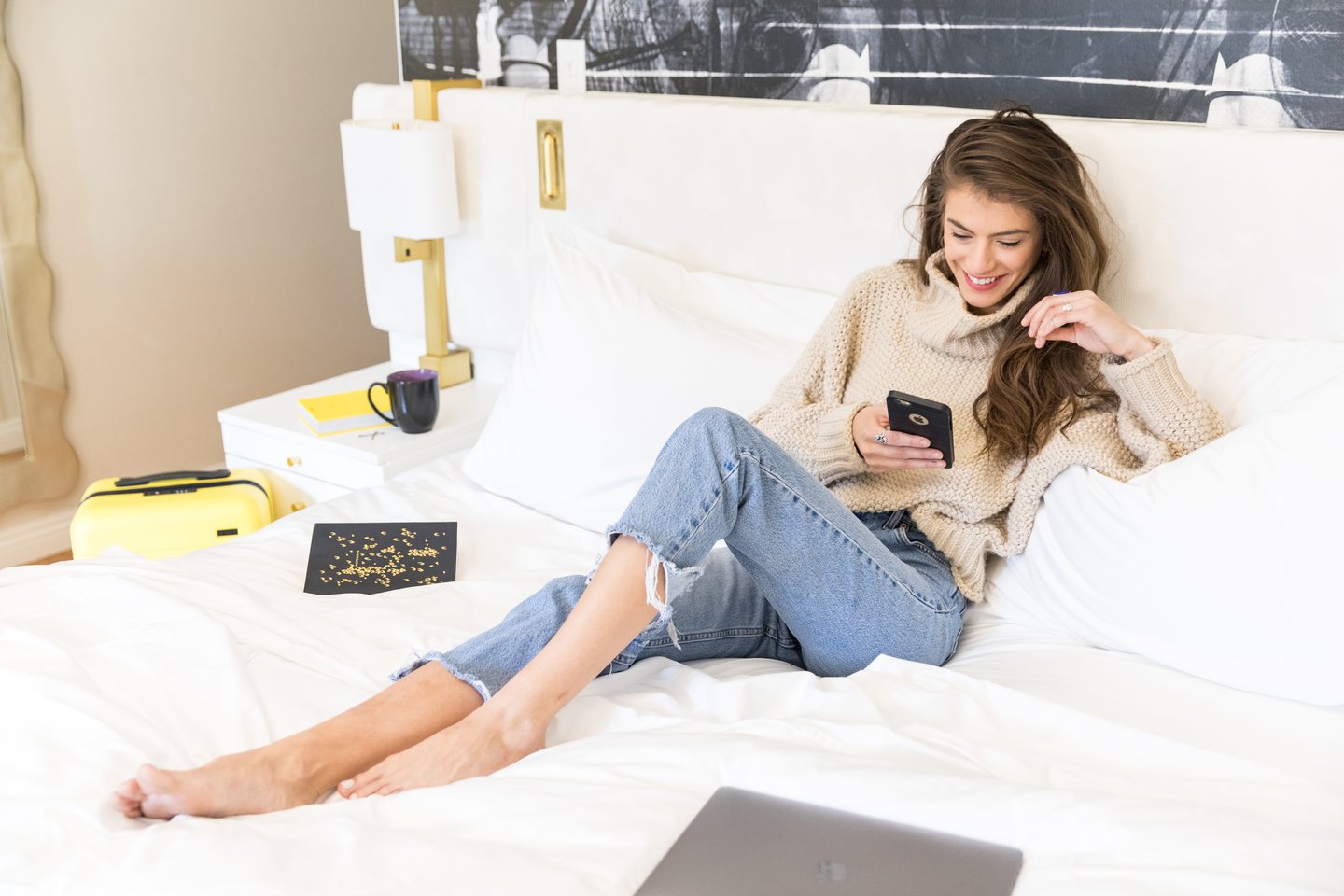 A woman in a beige sweater and ripped jeans sits on a white bed, smiling while looking at her phone. A mug, a notebook, and a closed laptop are nearby on the bed and nightstand.