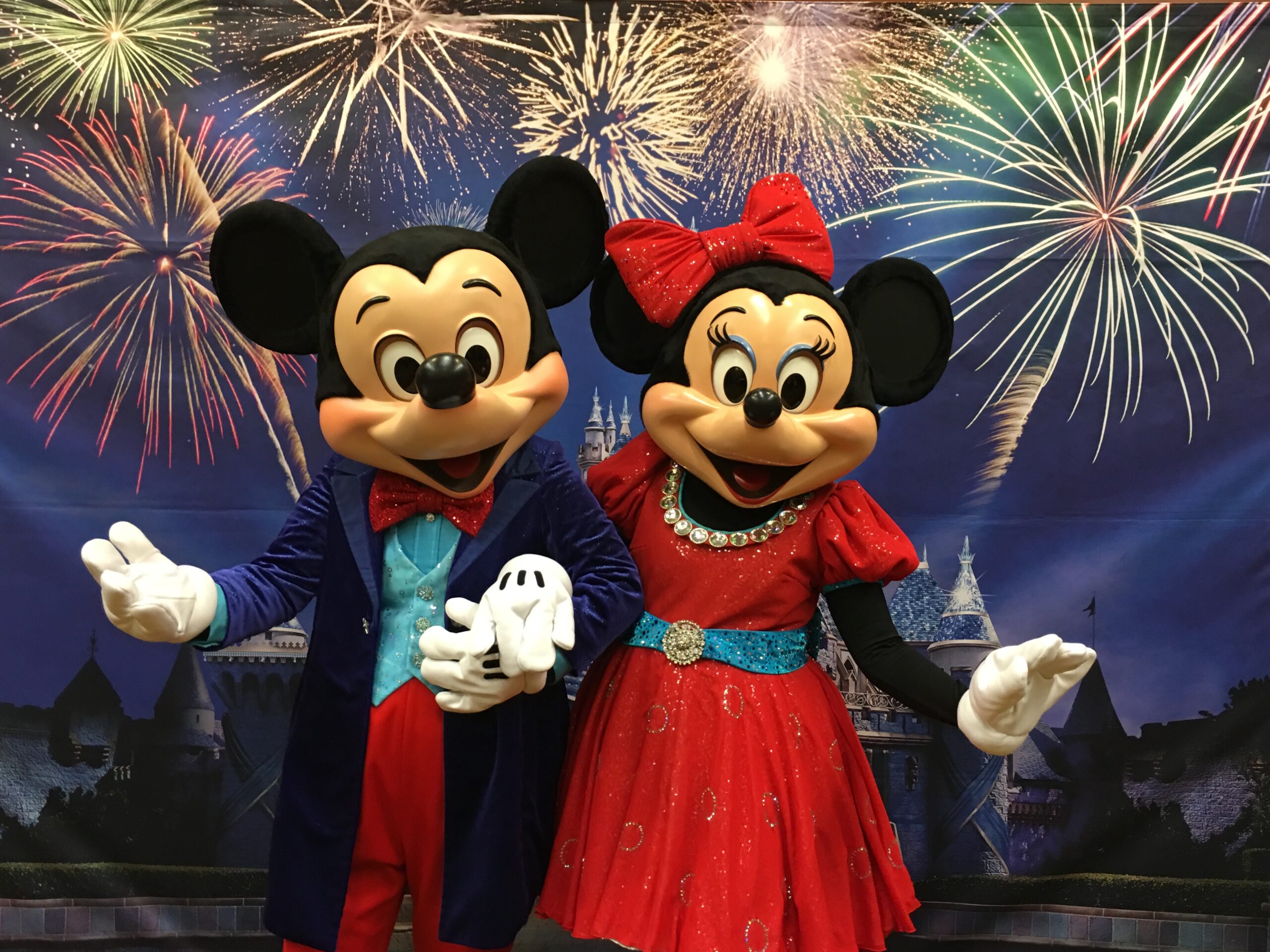 Mickey Mouse and Minnie Mouse, dressed in formal outfits, stand together smiling in front of a backdrop with colorful fireworks and a castle, capturing the magic often found at Costa Mesa attractions.