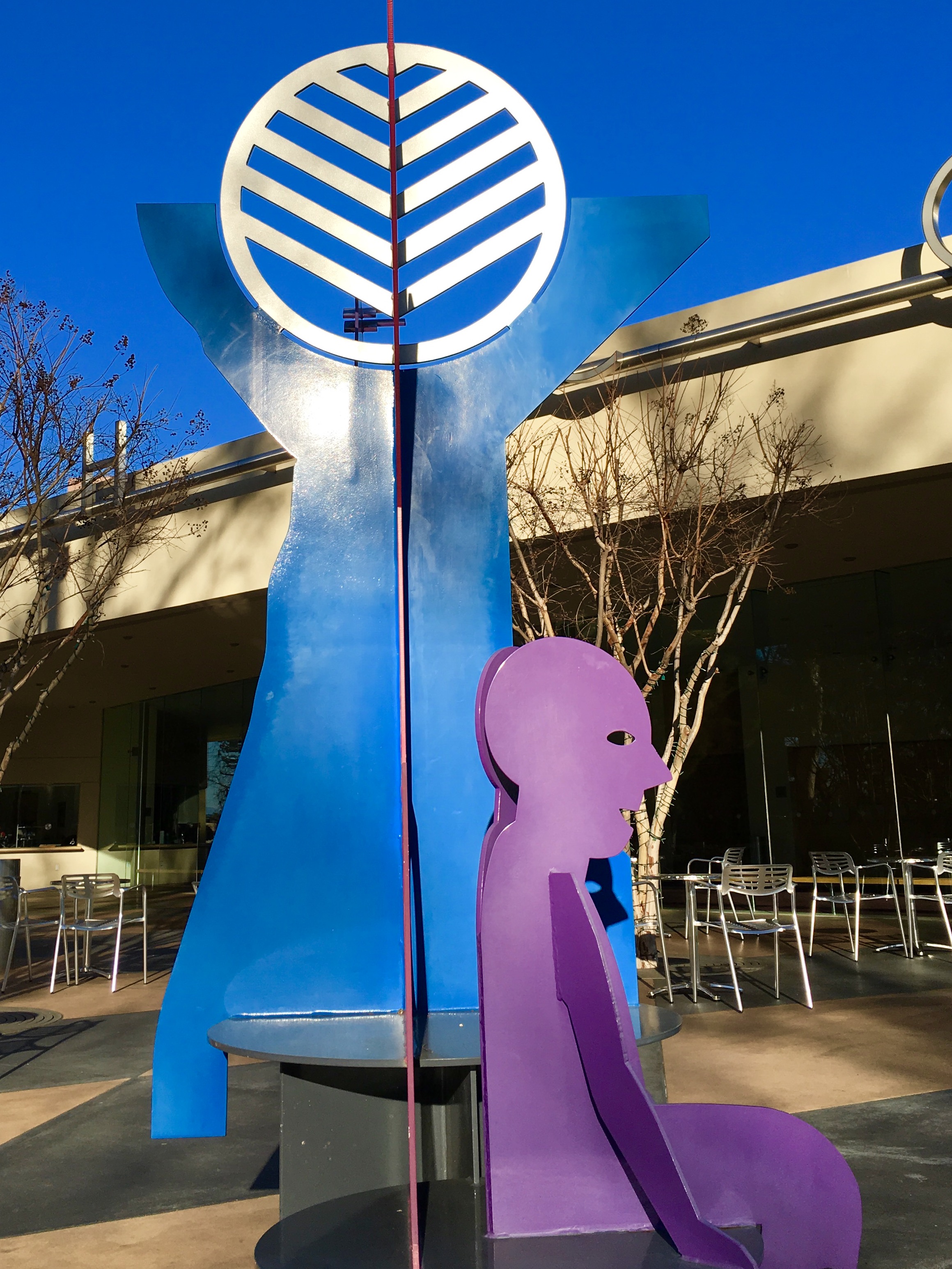 A colorful outdoor sculpture on the Costa Mesa Art Walk features a seated purple human silhouette before a tall blue figure with outstretched arms and a white circular design, set against a modern building and clear blue sky.