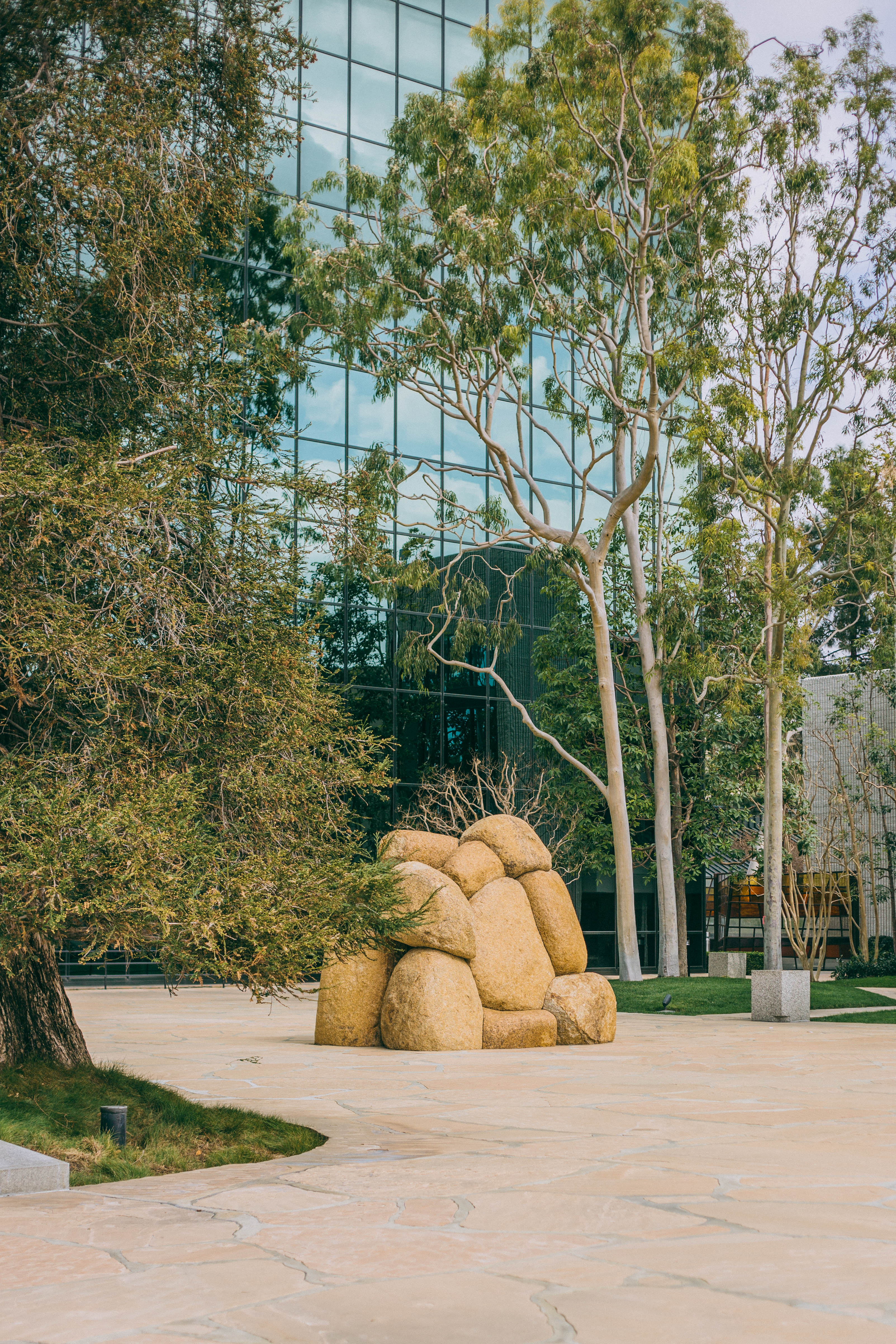 A modern glass building reflects trees in its facade. In the foreground, a cluster of large, rounded stone-like sculptures sits on a paved area, creating a striking scene along the Costa Mesa Art Walk amid lush greenery and tall trees.