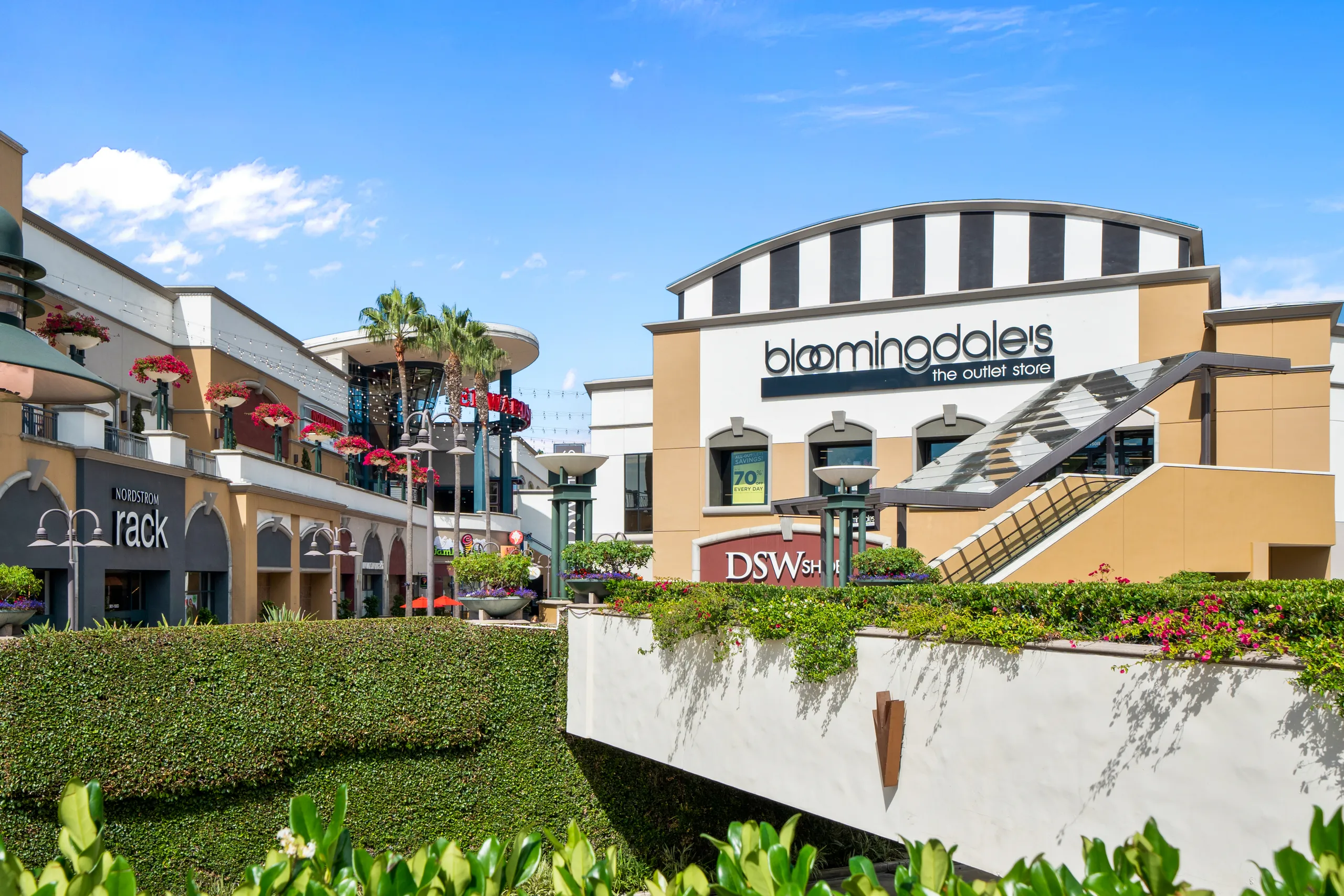 An outdoor shopping center in South Coast Metro, Costa Mesa, on a sunny day, featuring stores like Bloomingdale’s Outlet, Nordstrom Rack, and DSW amidst greenery, colorful flowers, and modern architecture.