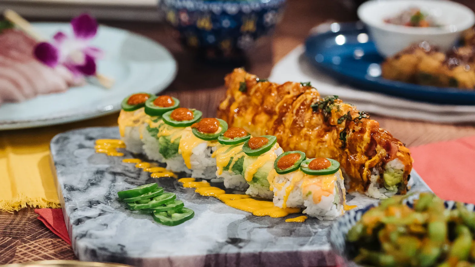A marble platter with sushi rolls topped with sliced jalapeños, orange sauce, and a reddish glaze, garnished with green chili peppers—just a taste of the best sushi Costa Mesa has to offer. Other dishes appear blurred in the background.
