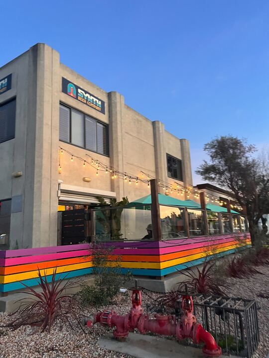 A modern two-story building with a Smith sign, colorful striped fencing, string lights, and an inviting outdoor seating area is landscaped with red plants and a fire hydrant—perfect for relaxing under clear blue skies at this Costa Mesa brewery.