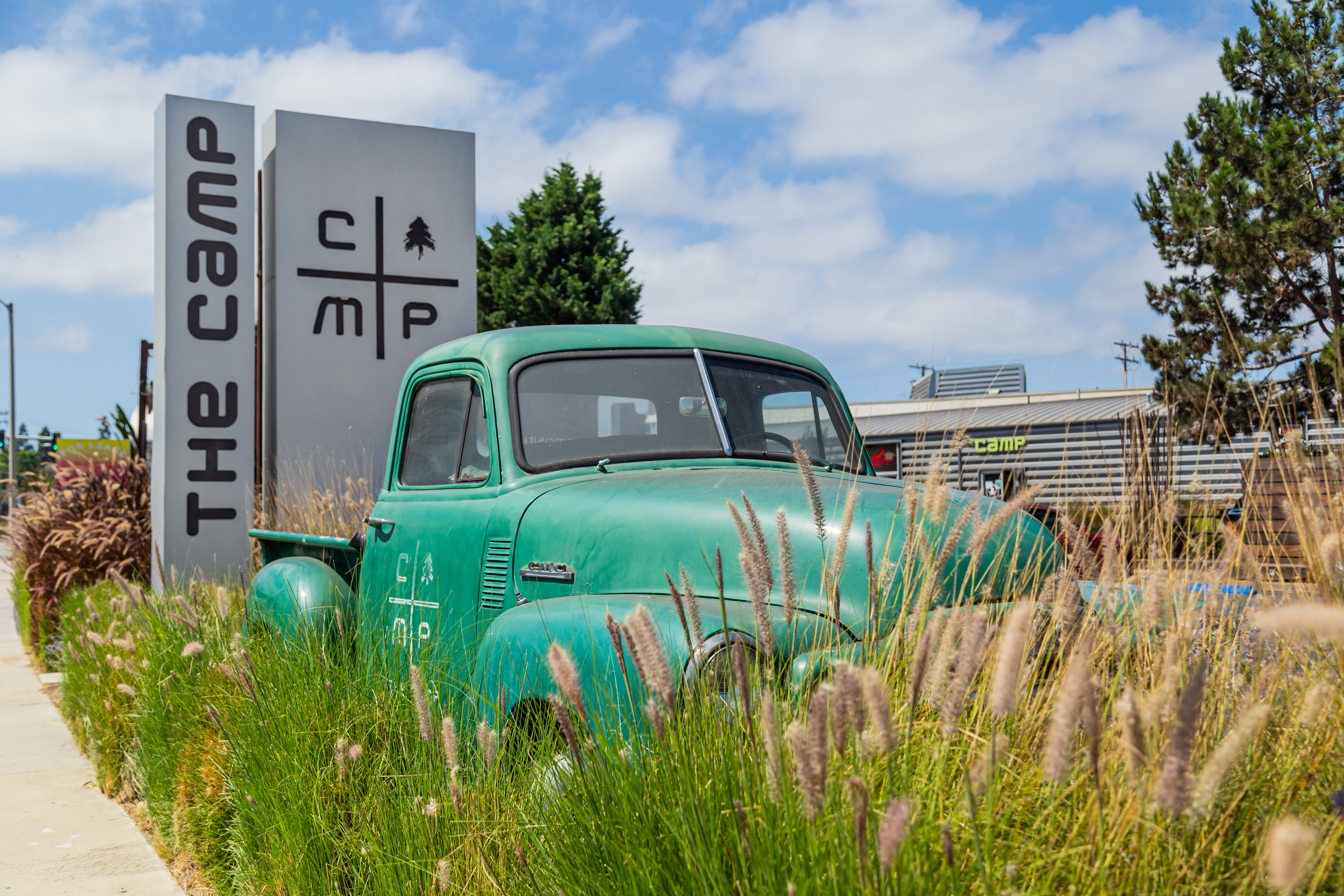 A vintage green truck is parked among tall grasses in front of a modern sign that reads The Camp and displays a logo, set in Costa Mesa, California, with trees and buildings in the background under a partly cloudy sky.