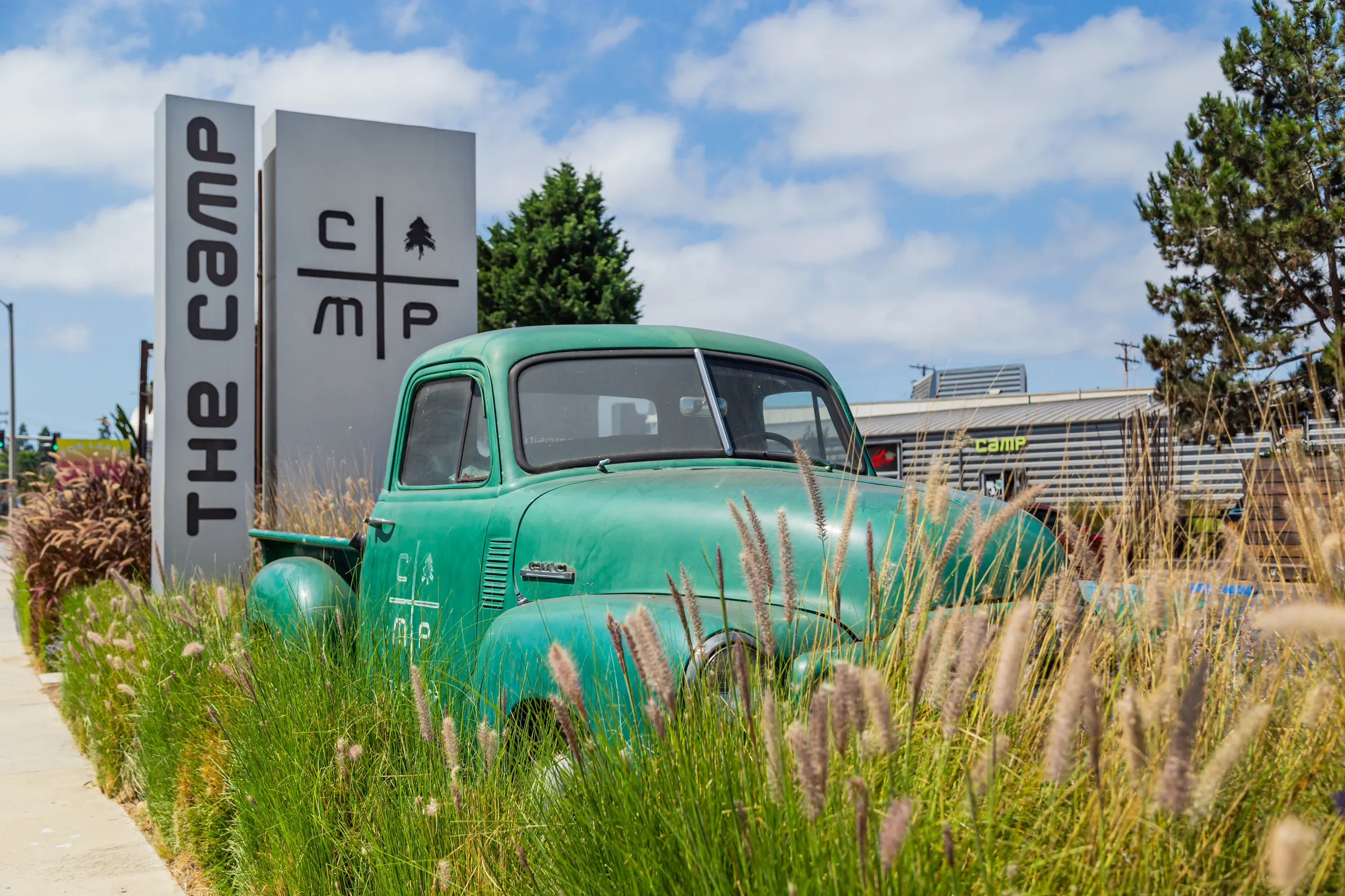 A vintage green truck is parked among tall grasses in front of a modern sign that reads The Camp and displays a logo, set in Costa Mesa, California, with trees and buildings in the background under a partly cloudy sky.