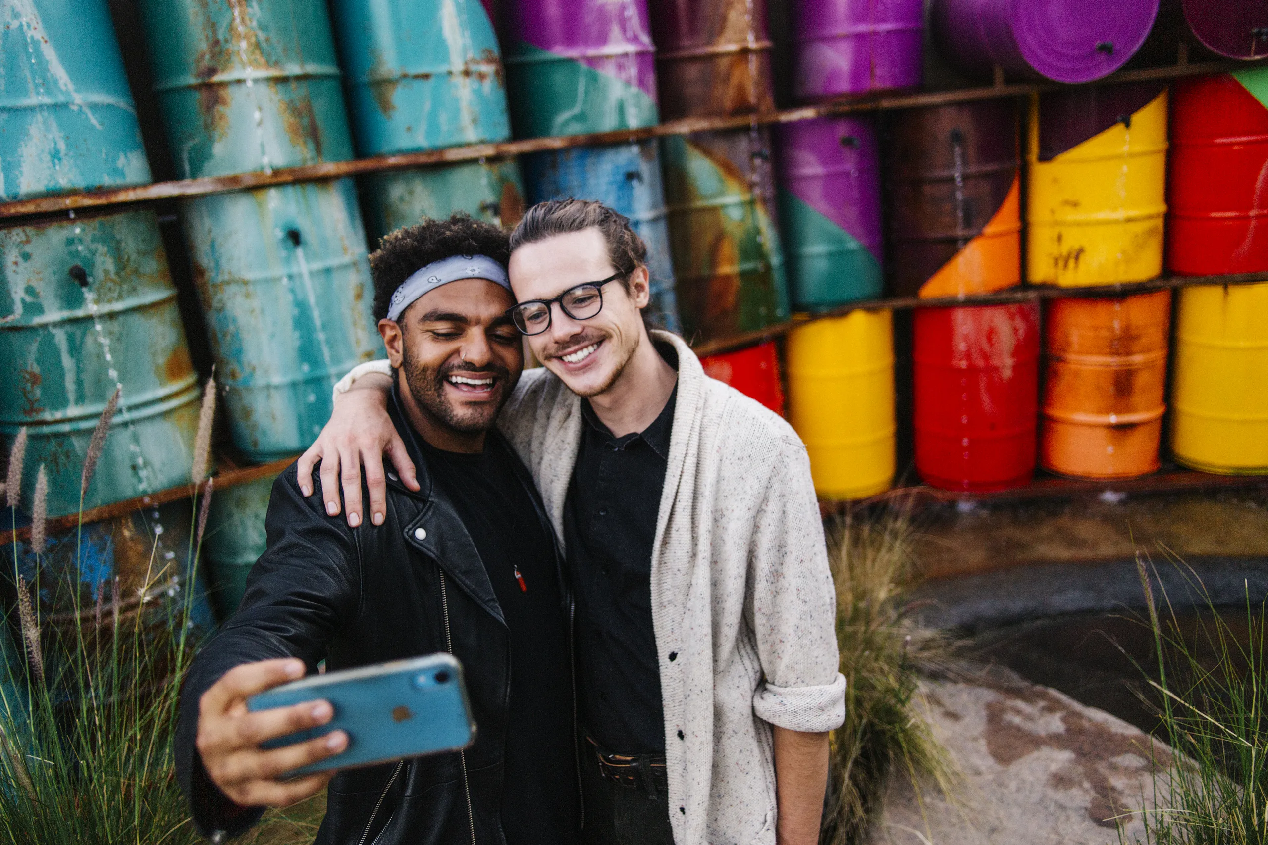 Two smiling friends take a selfie together in front of brightly colored, stacked barrels outdoors in Costa Mesa, California. One has a headband and leather jacket, while the other wears glasses and a cardigan.