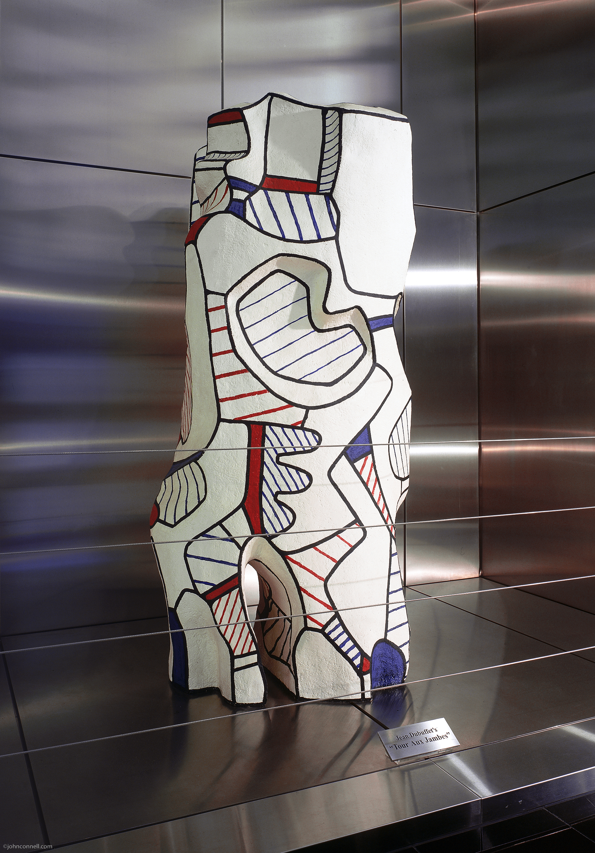 Abstract sculpture with bold black outlines and geometric shapes in white, red, and blue stands before reflective metal panels—a highlight of the Costa Mesa Art Walk. A small plaque is visible at the base of the artwork.