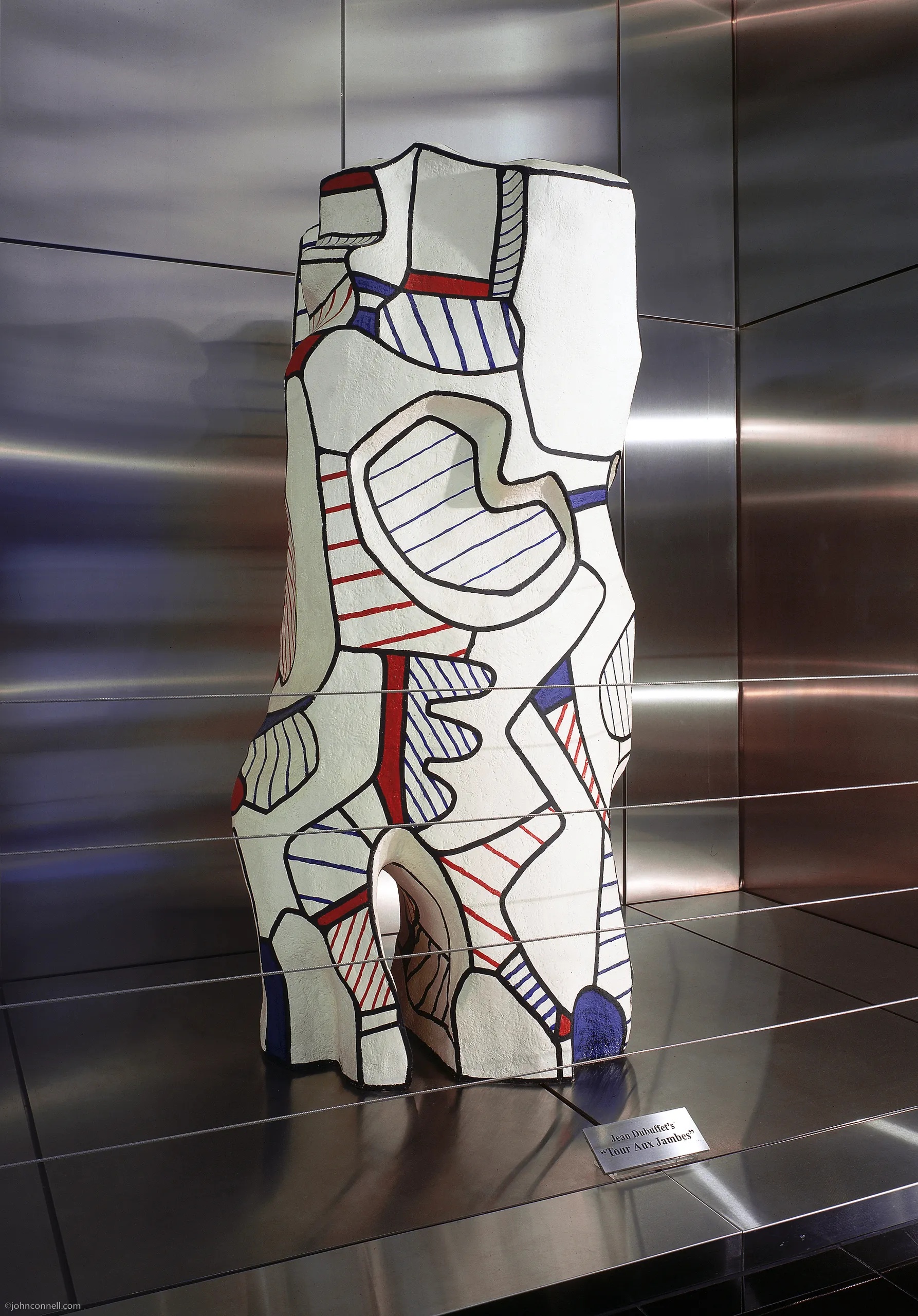 Abstract sculpture with bold black outlines and geometric shapes in white, red, and blue stands before reflective metal panels—a highlight of the Costa Mesa Art Walk. A small plaque is visible at the base of the artwork.