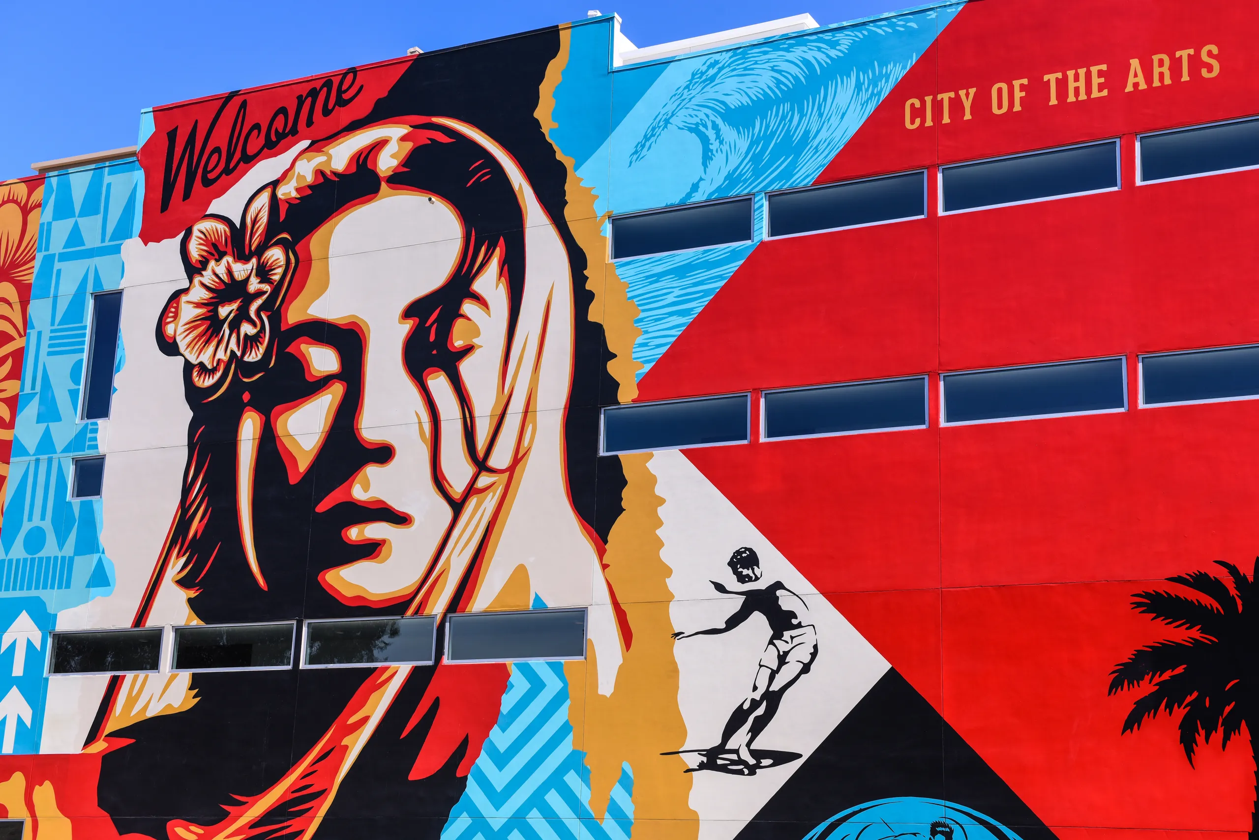 Colorful mural on a building in Costa Mesa, California, showing a woman's face with a flower in her hair, a surfer, palm tree, and bold geometric shapes. The words Welcome and City of the Arts are painted on the upper part.