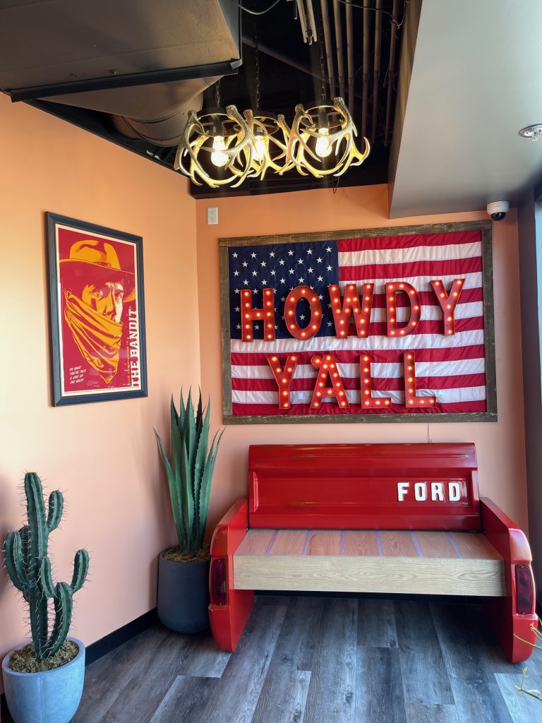 A room with an American flag, lit “Howdy Y’all” sign, red Ford bench, two potted cacti, a framed cowboy poster, and an antler chandelier on a peach wall.