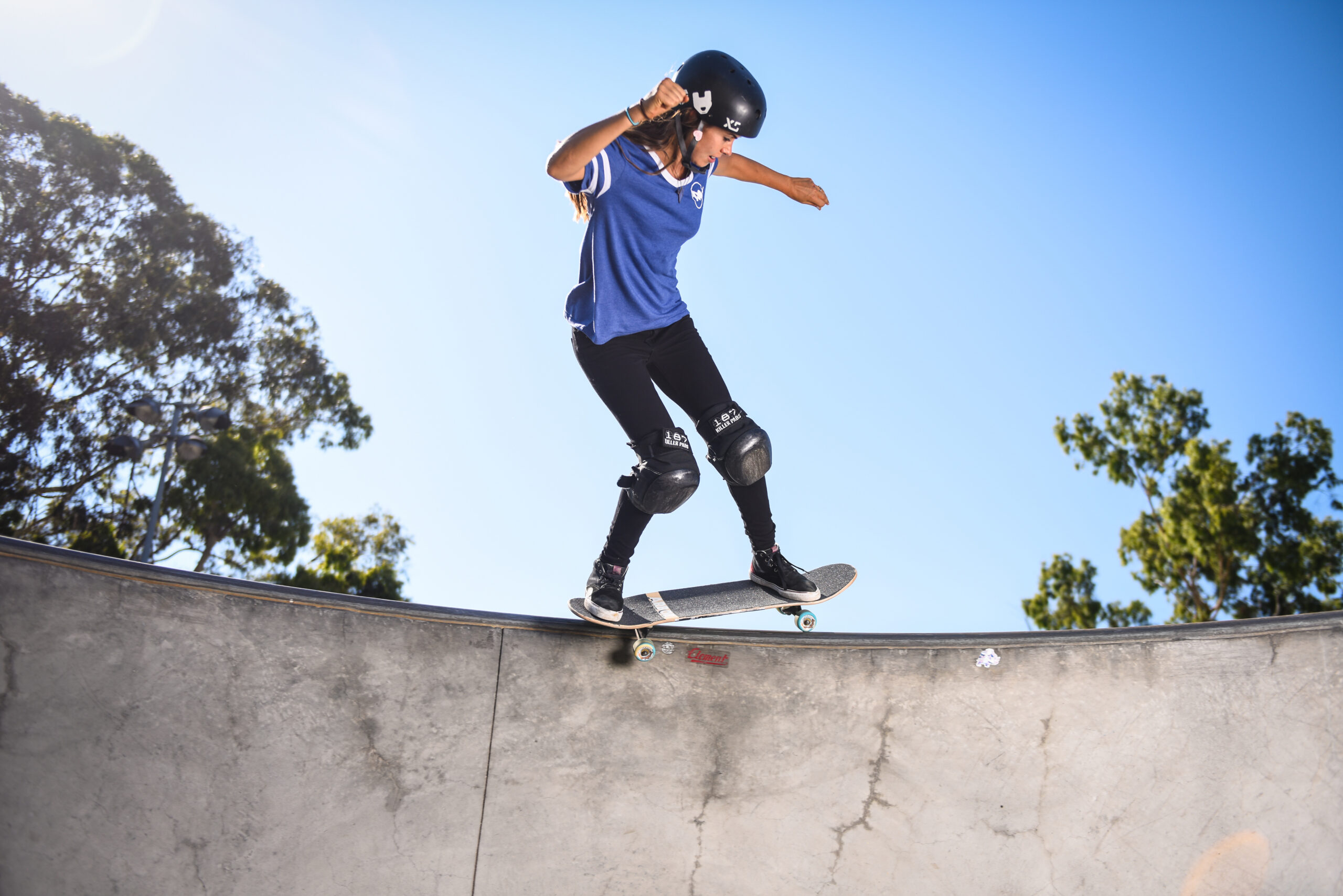A skateboarder wearing a helmet and knee pads balances on the edge of a concrete skatepark ramp under a clear blue sky, surrounded by green trees, much like you’d find in Costa Mesa.