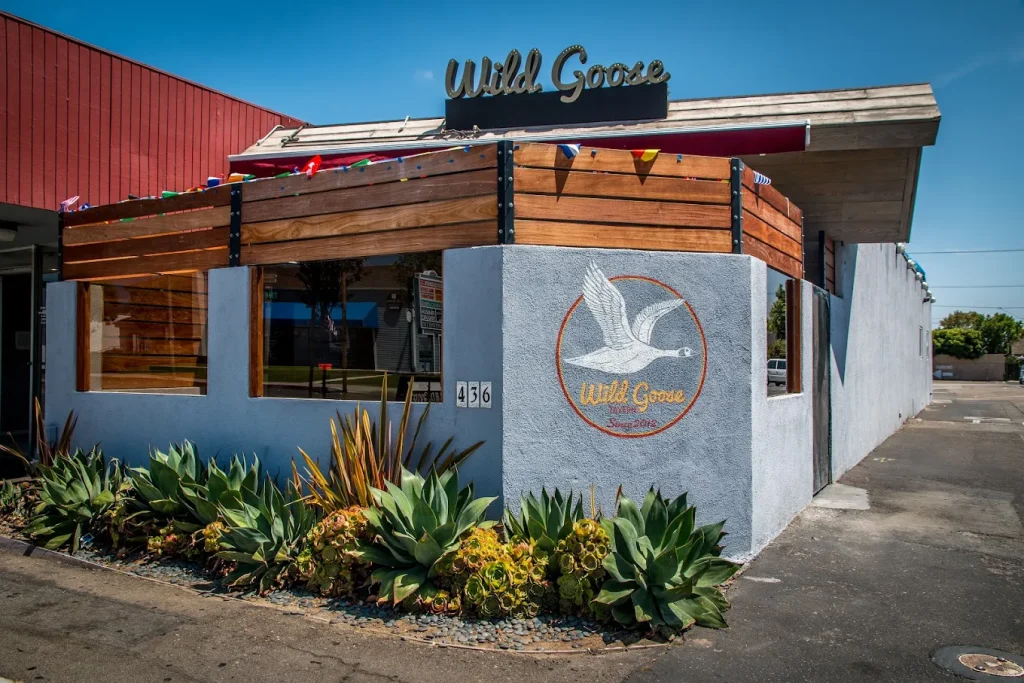 A corner view of the Wild Goose restaurant, featuring a mural of a flying goose on a light blue wall, wooden paneling, and green succulents planted along the sidewalk under a sunny sky.