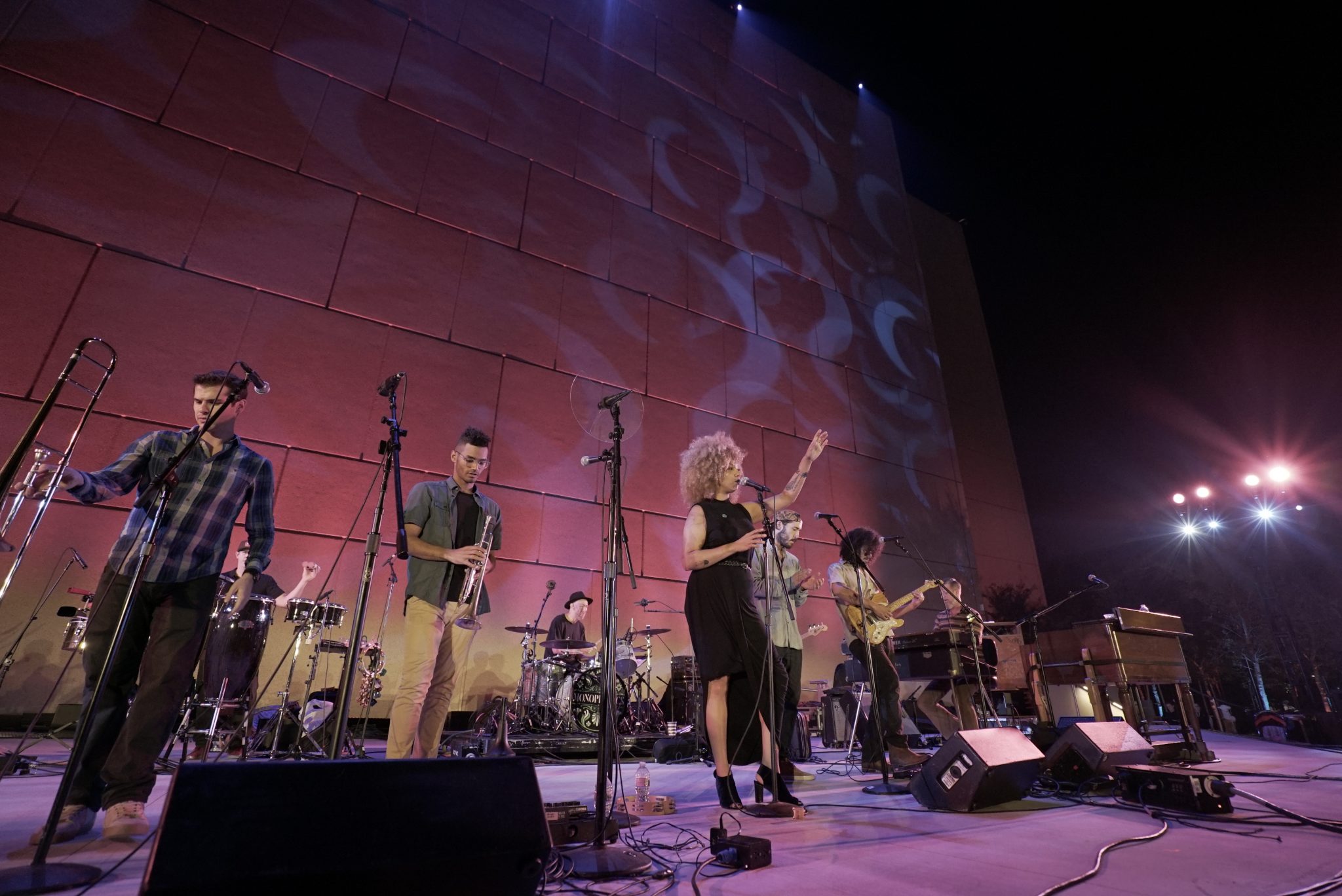 A band performs on an outdoor stage at night, with multiple musicians playing instruments and a woman singing into a microphone. The background features a tall wall lit by purple stage lights.