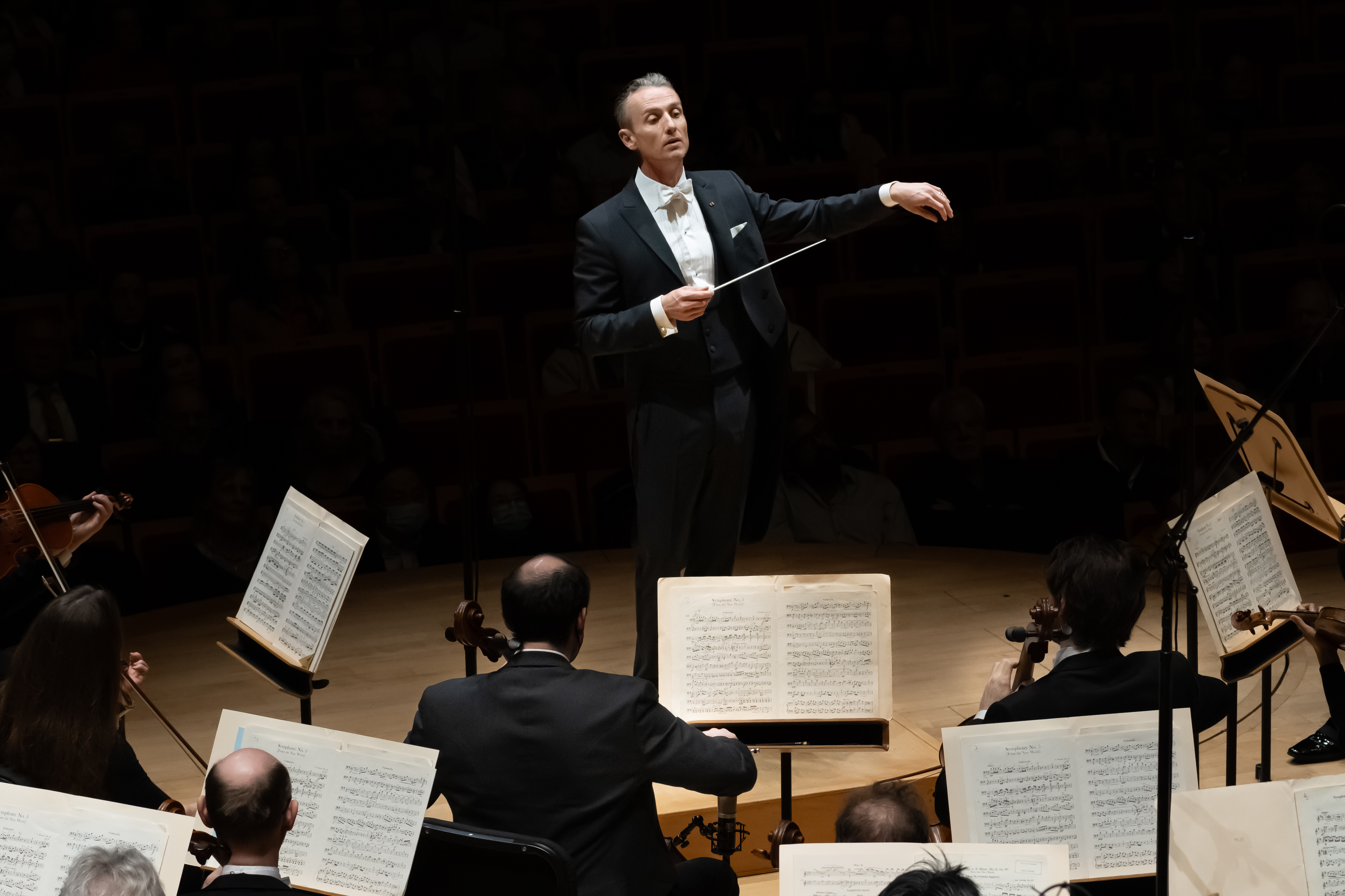 A conductor in a tuxedo stands on stage at the Segerstrom Performing Arts Center Costa Mesa, leading an orchestra. Musicians with sheet music sit before him, while the dimly lit concert hall hosts an attentive audience in the background.