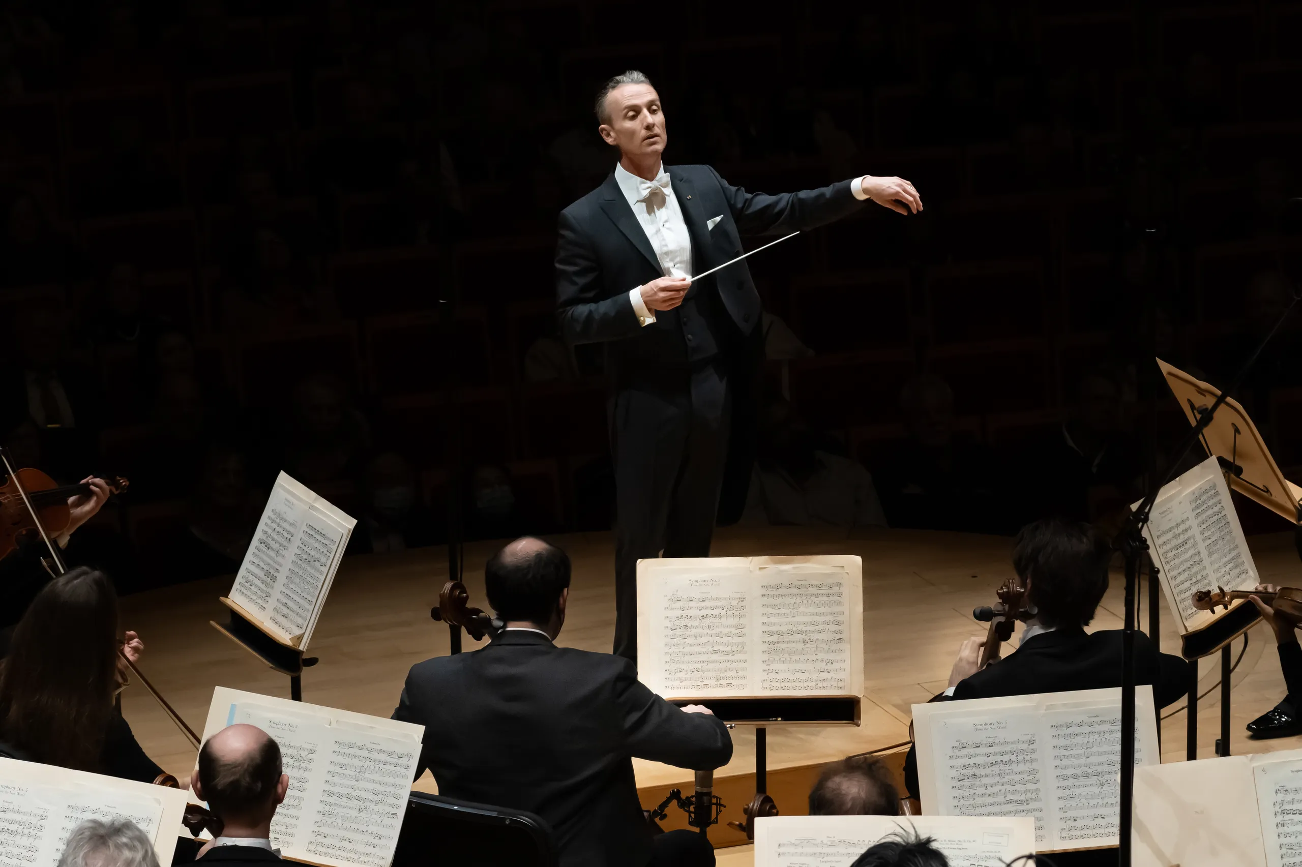 A conductor in a tuxedo stands on stage at the Segerstrom Performing Arts Center Costa Mesa, leading an orchestra. Musicians with sheet music sit before him, while the dimly lit concert hall hosts an attentive audience in the background.