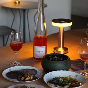 A dimly lit table set with wine glasses, a tall bottle of rosé wine, elegant dishes of gourmet food, and a small modern lamp creating a cozy dining atmosphere—perfect for those exploring things to do in Costa Mesa.