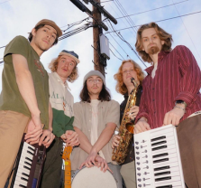 Five young men stand outdoors under power lines, each holding musical instruments including keyboards, a guitar, a saxophone, and a drum. They look towards the camera with relaxed expressions.