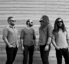 Four men with beards and sunglasses stand in front of a wooden wall, casually dressed and looking at each other in a relaxed, informal group pose.